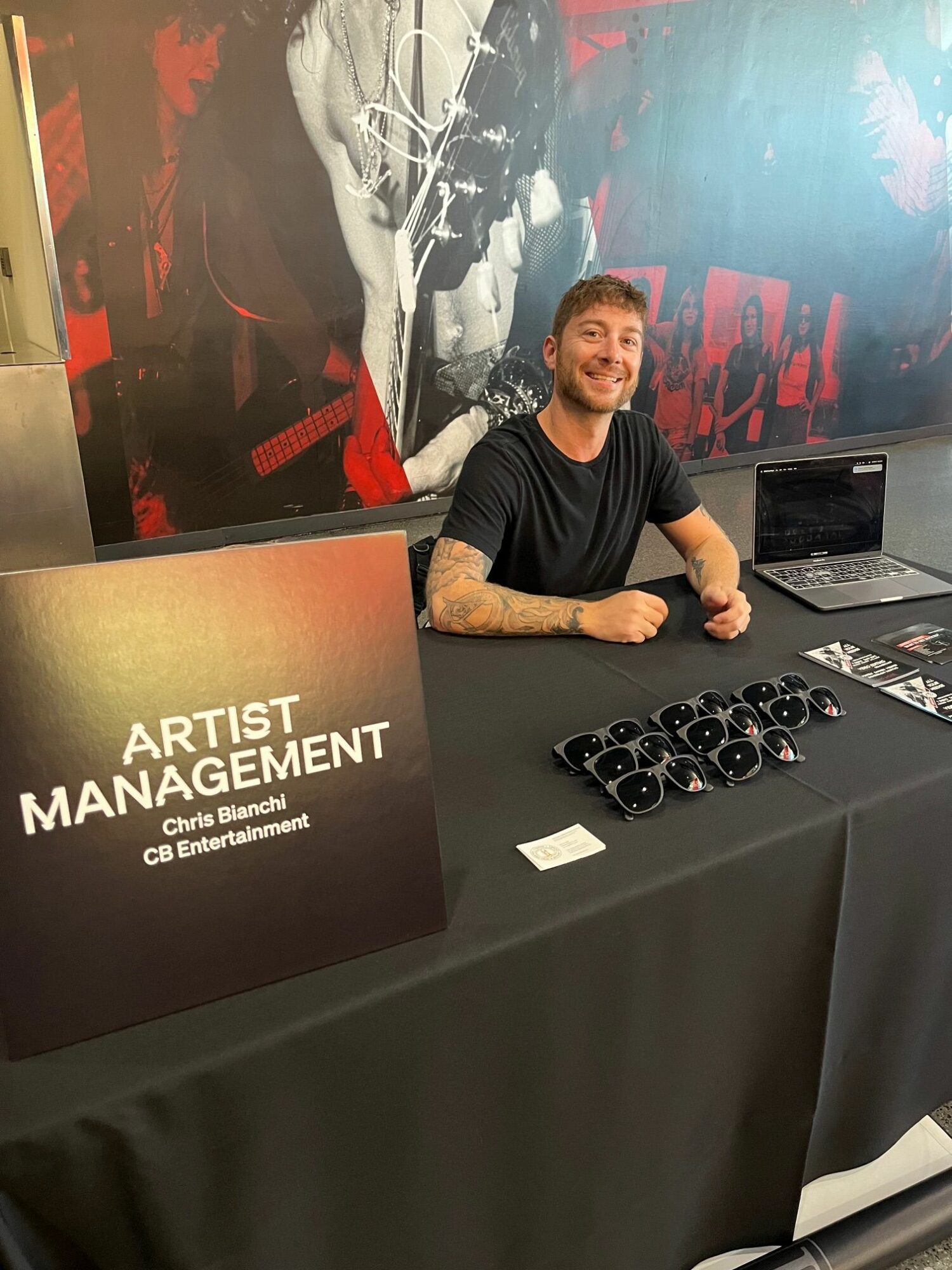 Man sitting at a table with a sign reading 'ARTIST MANAGEMENT' and a laptop, smiling at the camera.