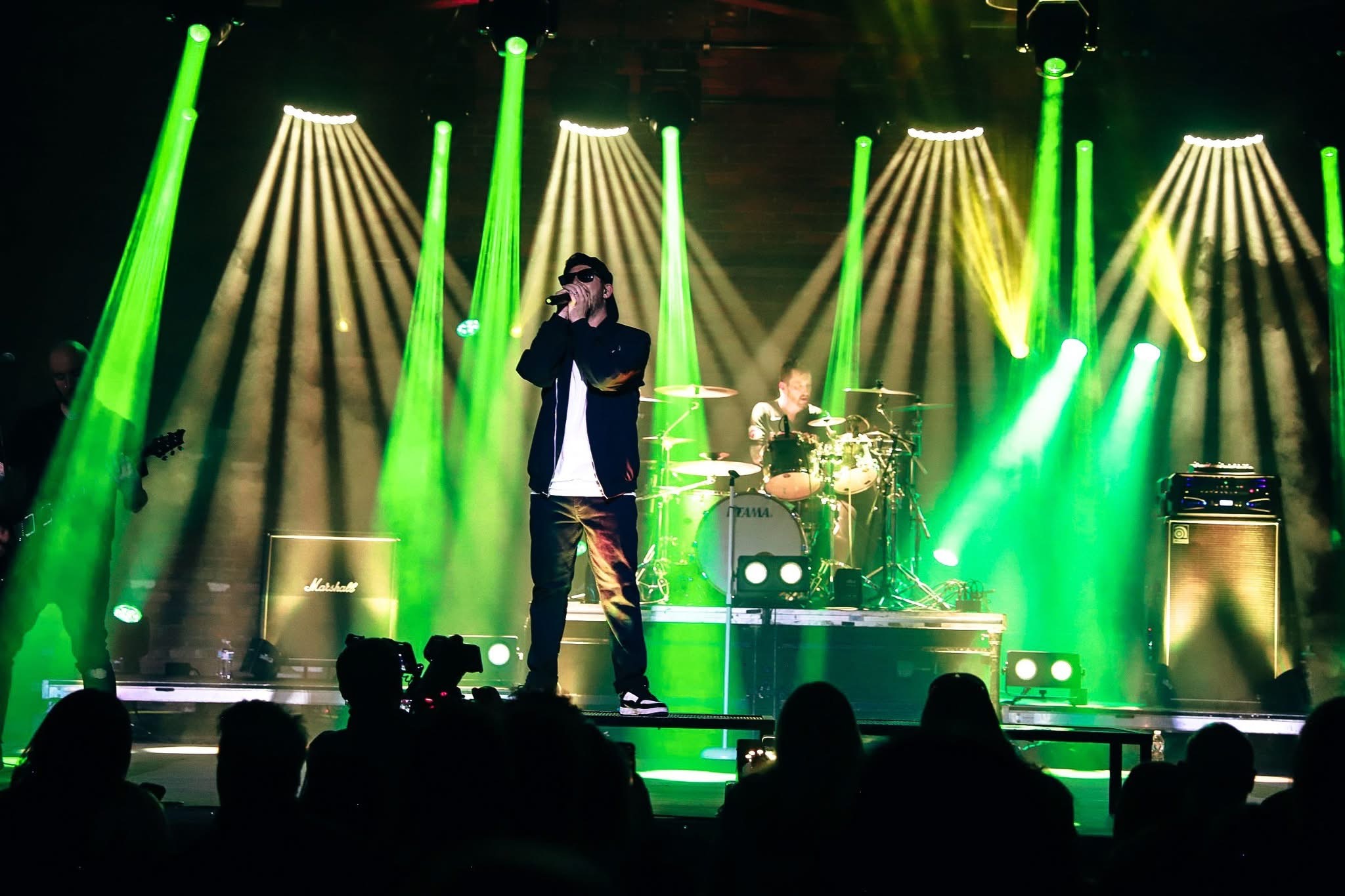 Performer singing on stage with colorful green and yellow lights, drums, and amplifiers in the background.