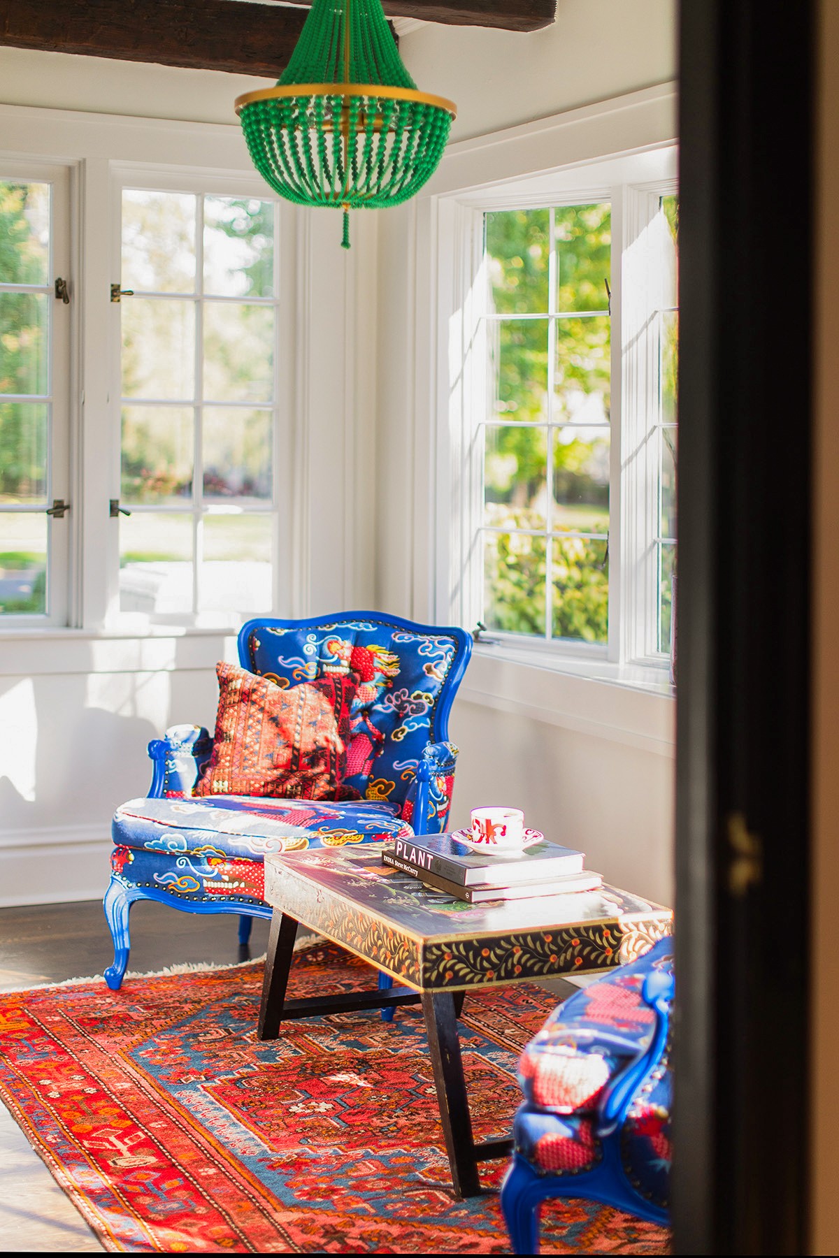 Bright room with large windows, blue armchair, colorful pillows, and a patterned table on a red rug.