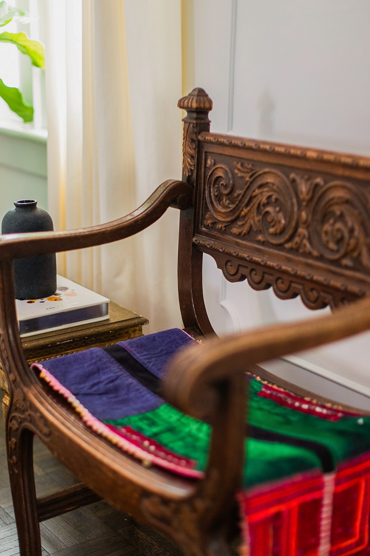 Close-up of an ornate wooden chair with colorful fabric on the seat, a vase on a side table, and curtains in the background.
