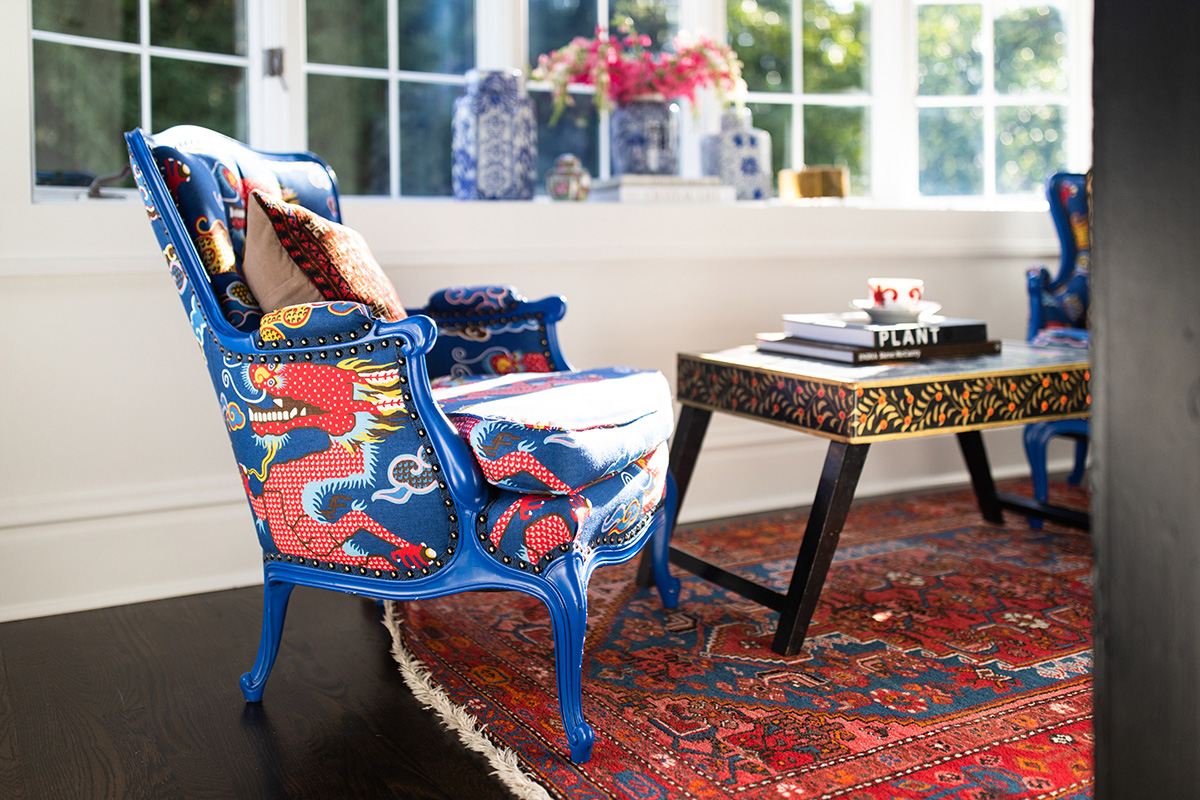 Blue armchair with patterned fabric and a pillow, next to a wooden table with books, in a room with large windows and a rug.