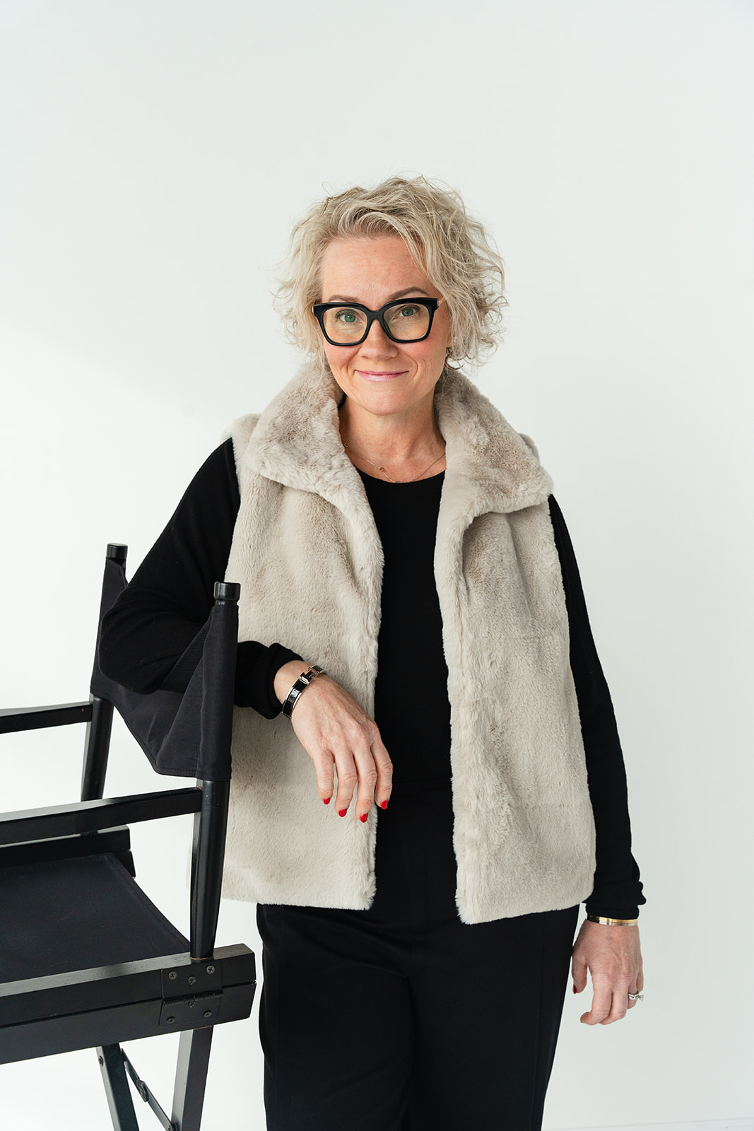Older woman with curly gray hair and glasses standing next to a black chair, wearing a black outfit and a light-colored vest.