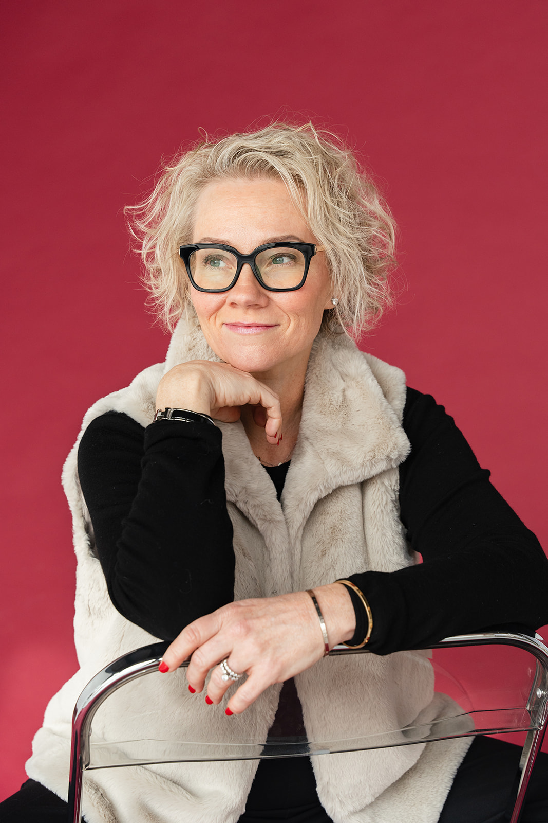 Woman with curly gray hair and glasses resting her chin on her hand, smiling, against a pink background.