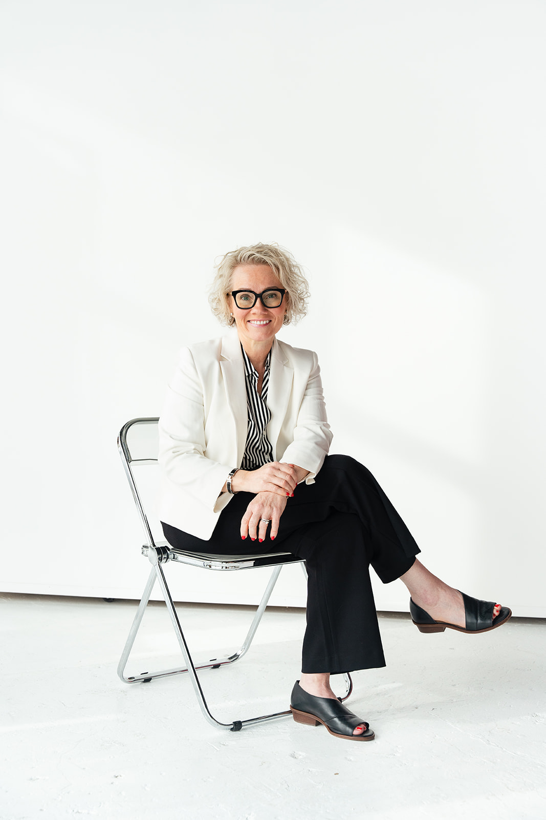 Woman with curly gray hair and glasses sitting on a folding chair, smiling, wearing a white blazer and black pants.