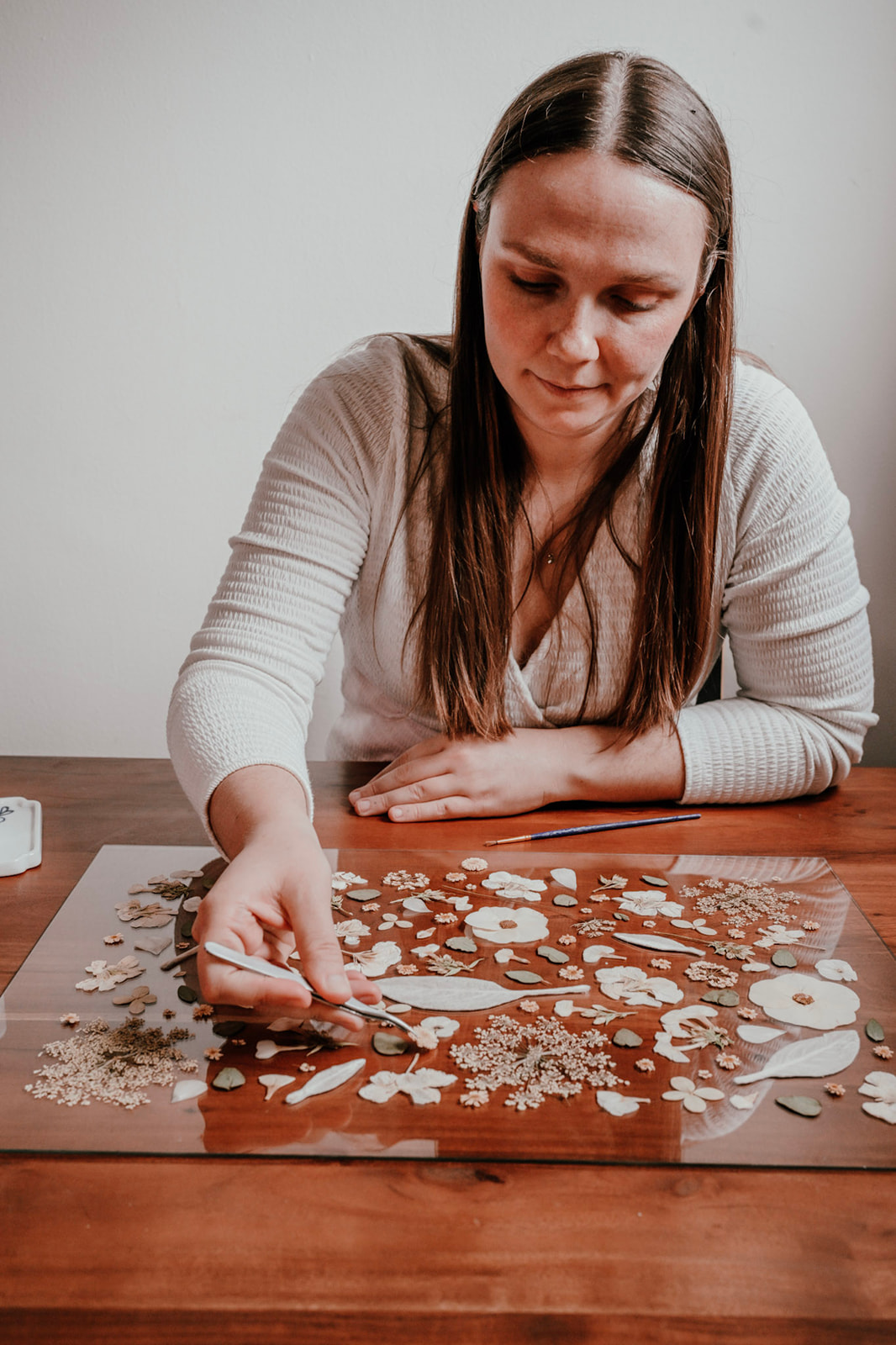 Woman with long brown hair arranging puzzle pieces on a wooden table, wearing a white long-sleeve shirt.