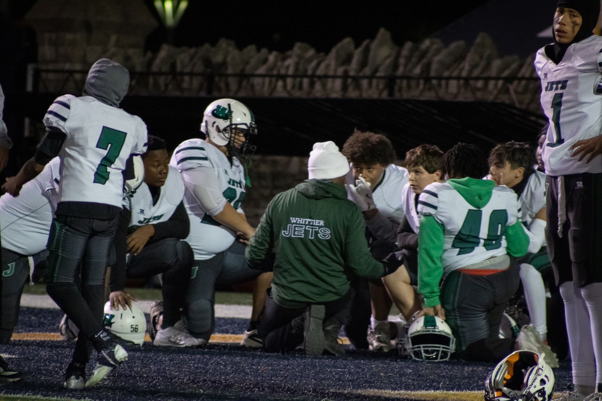 Football team huddled on field with coach, players wearing white and green uniforms, helmet on ground, stadium background.