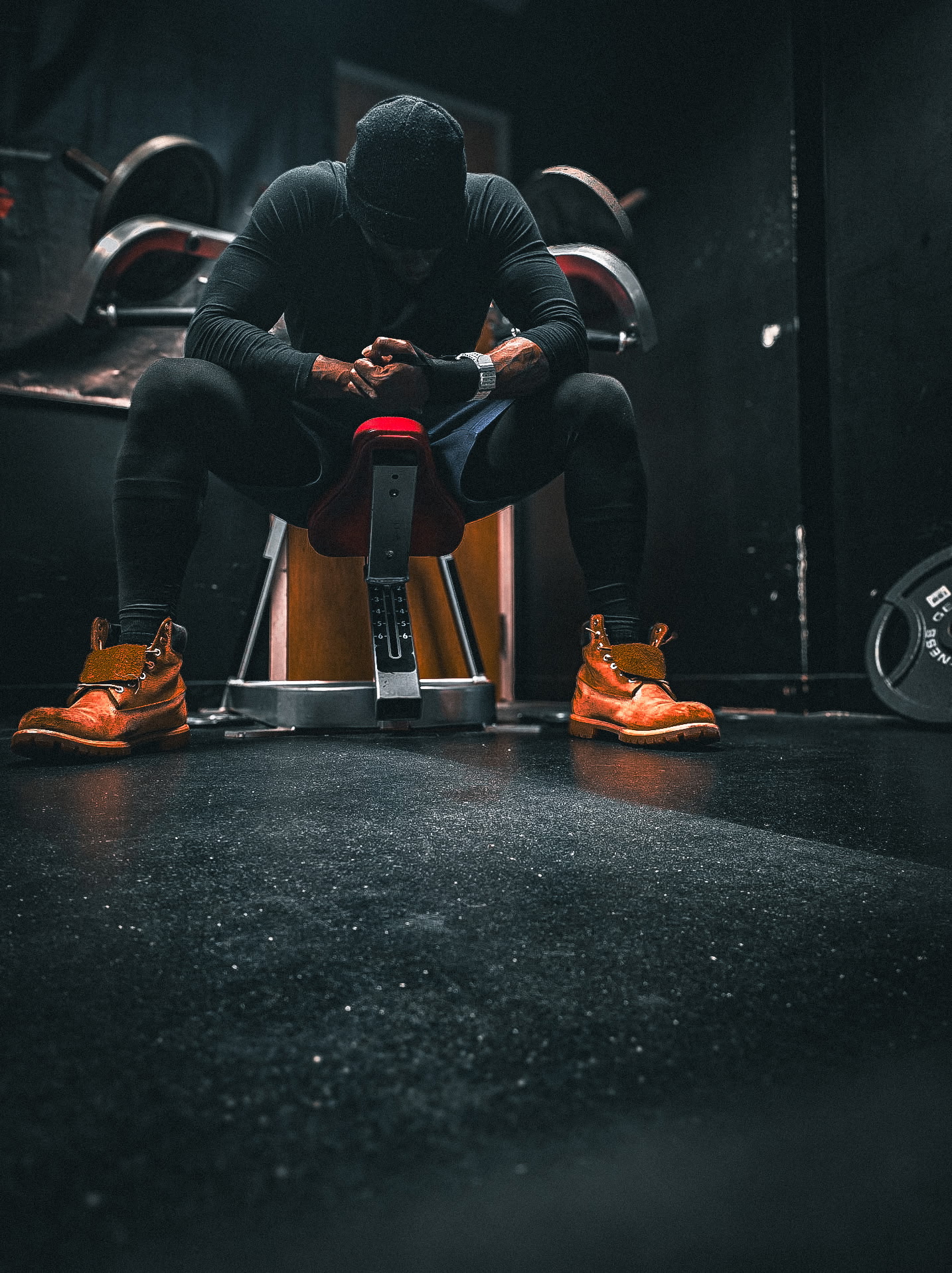 Person sitting on a bench in a gym, wearing black clothing and orange sneakers, with gym equipment in background.