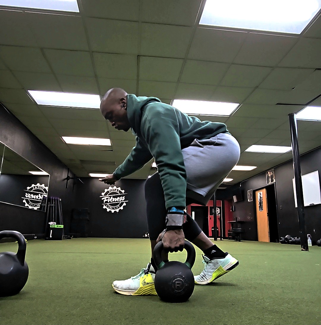 Man in gym lifting kettlebell with both hands, bent over, in a workout room with green floor and ceiling lights.