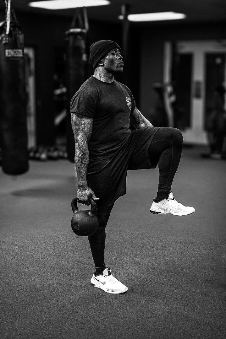 Person lifting a kettlebell in a gym, wearing a black shirt, shorts, and white sneakers.