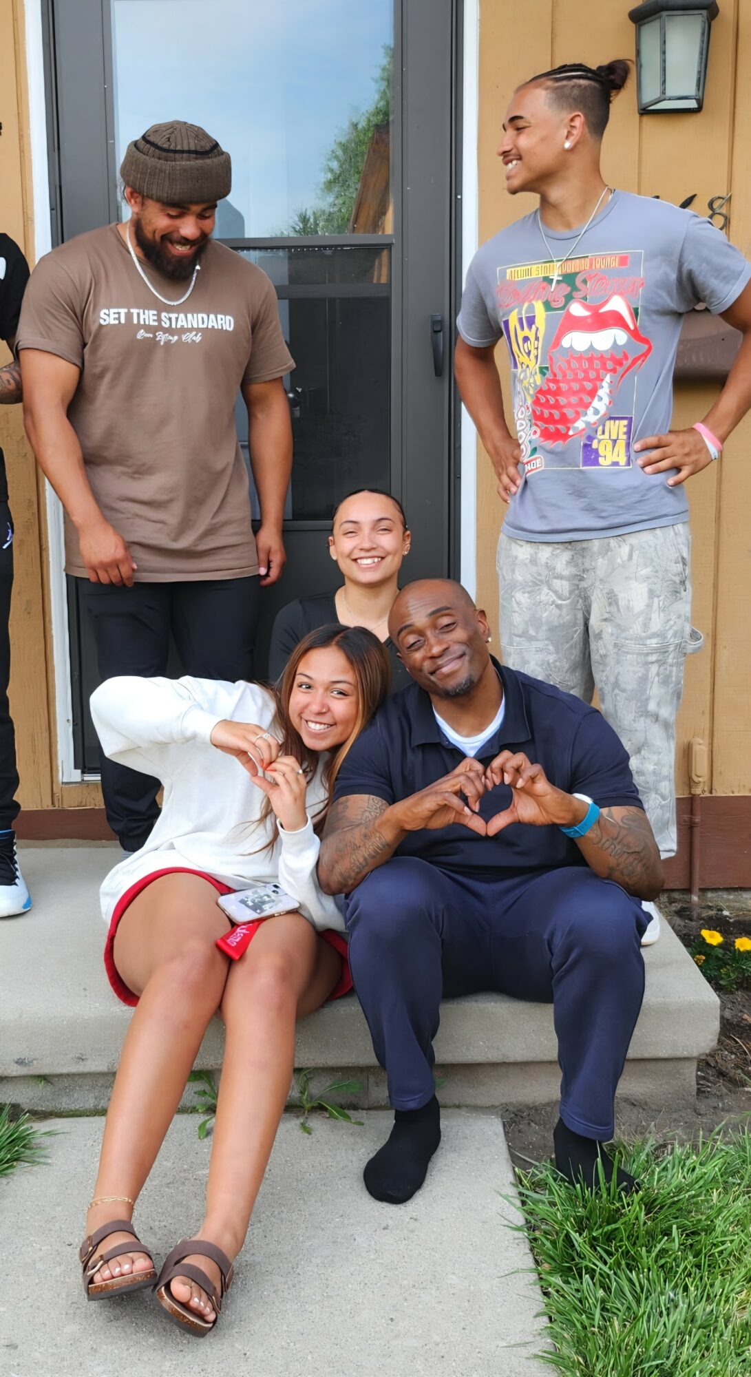 Group of five people sitting and standing on porch steps outside a house, smiling and interacting.