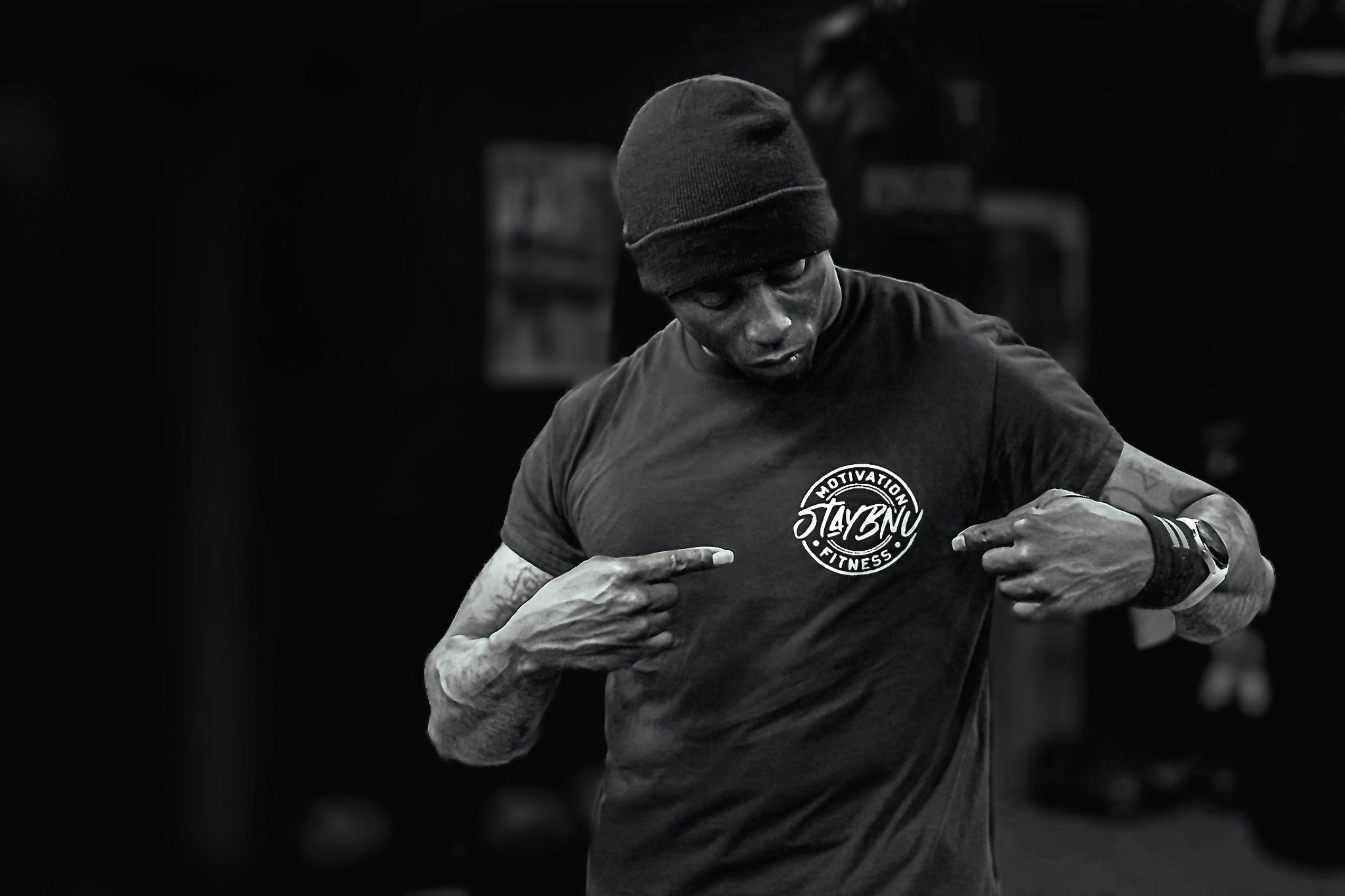 Man wearing a beanie and T-shirt pointing at his chest in a gym setting.