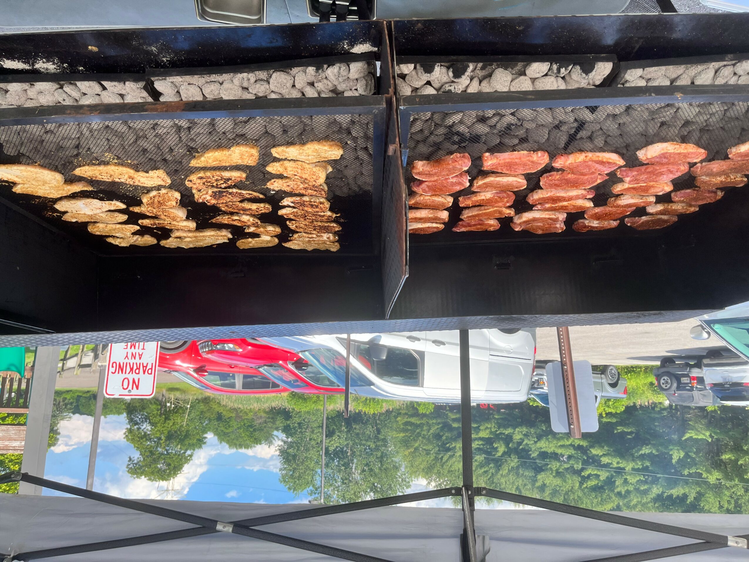 Two black grills with chicken and sausage, viewed upside down, with cars and trees in the background.