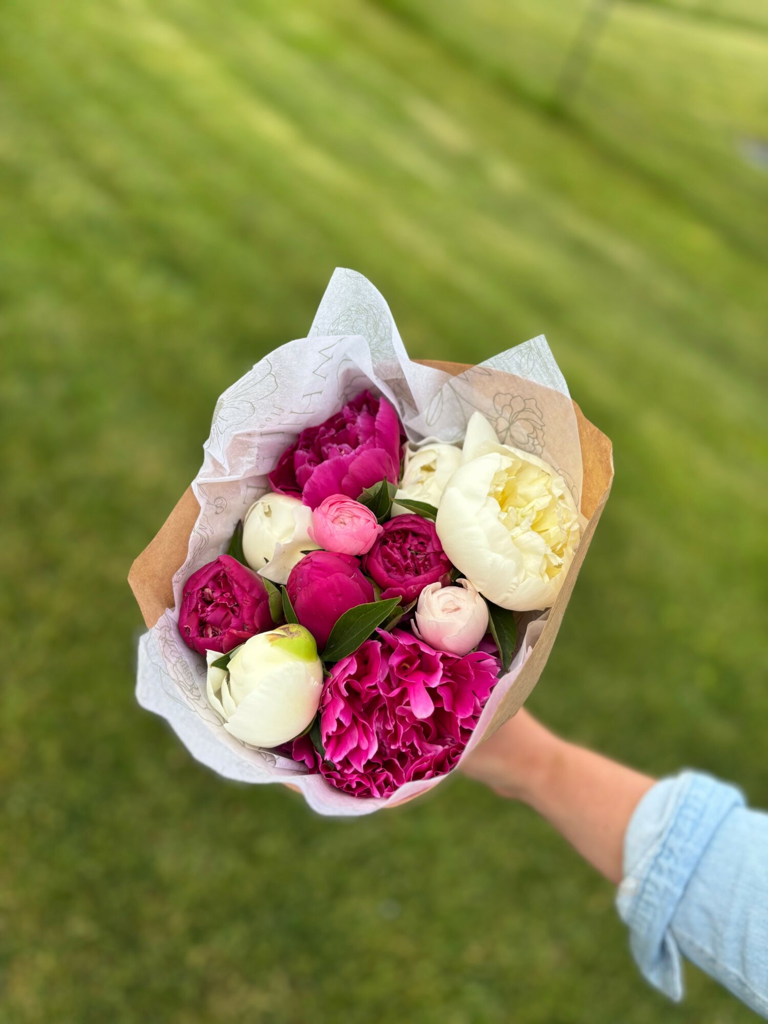 Person holding a bouquet of pink and white flowers wrapped in paper outdoors on grass.