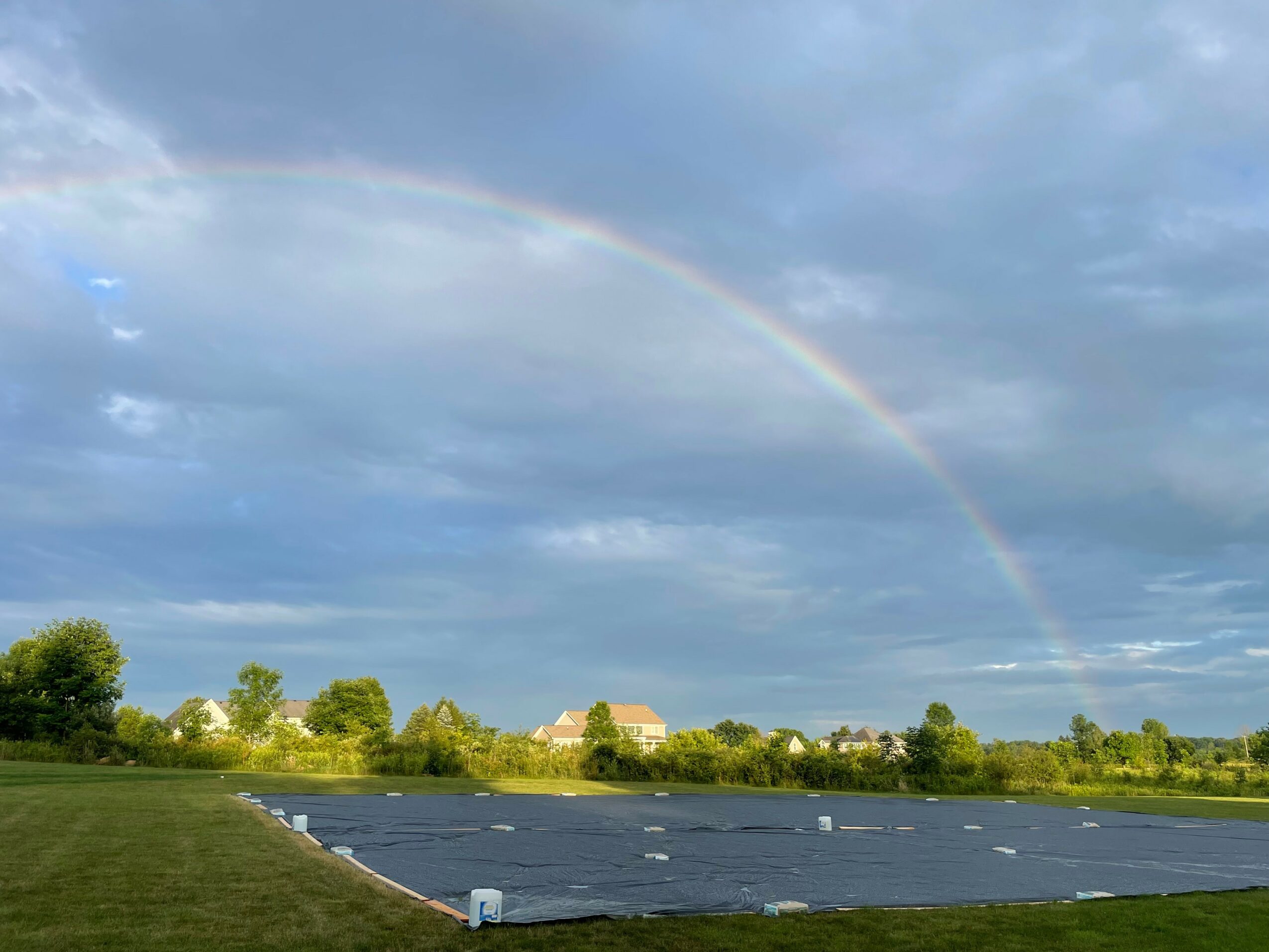 Rainbow arching over a cloudy sky above a landscape with trees and a parking lot.
