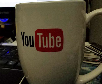 White mug with YouTube logo and text, placed on a desk with papers and a keyboard nearby.