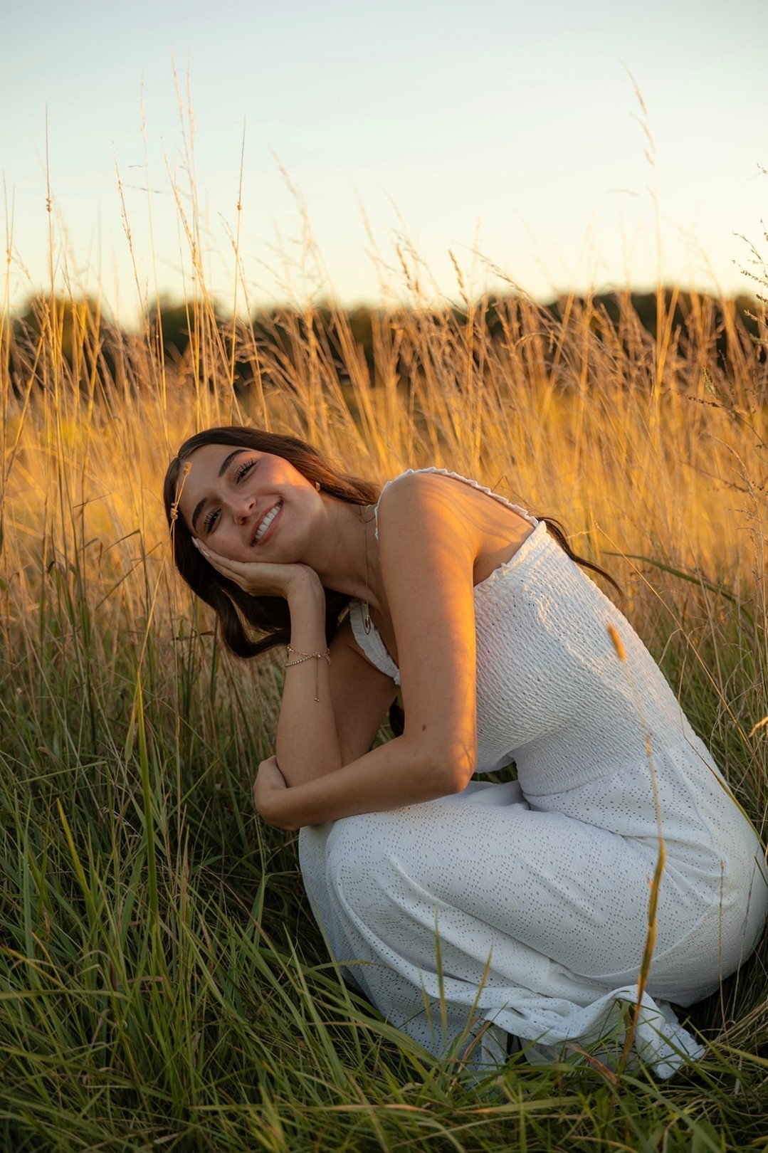 Young woman in white dress crouching in tall grass, smiling, with sunset sky in background.
