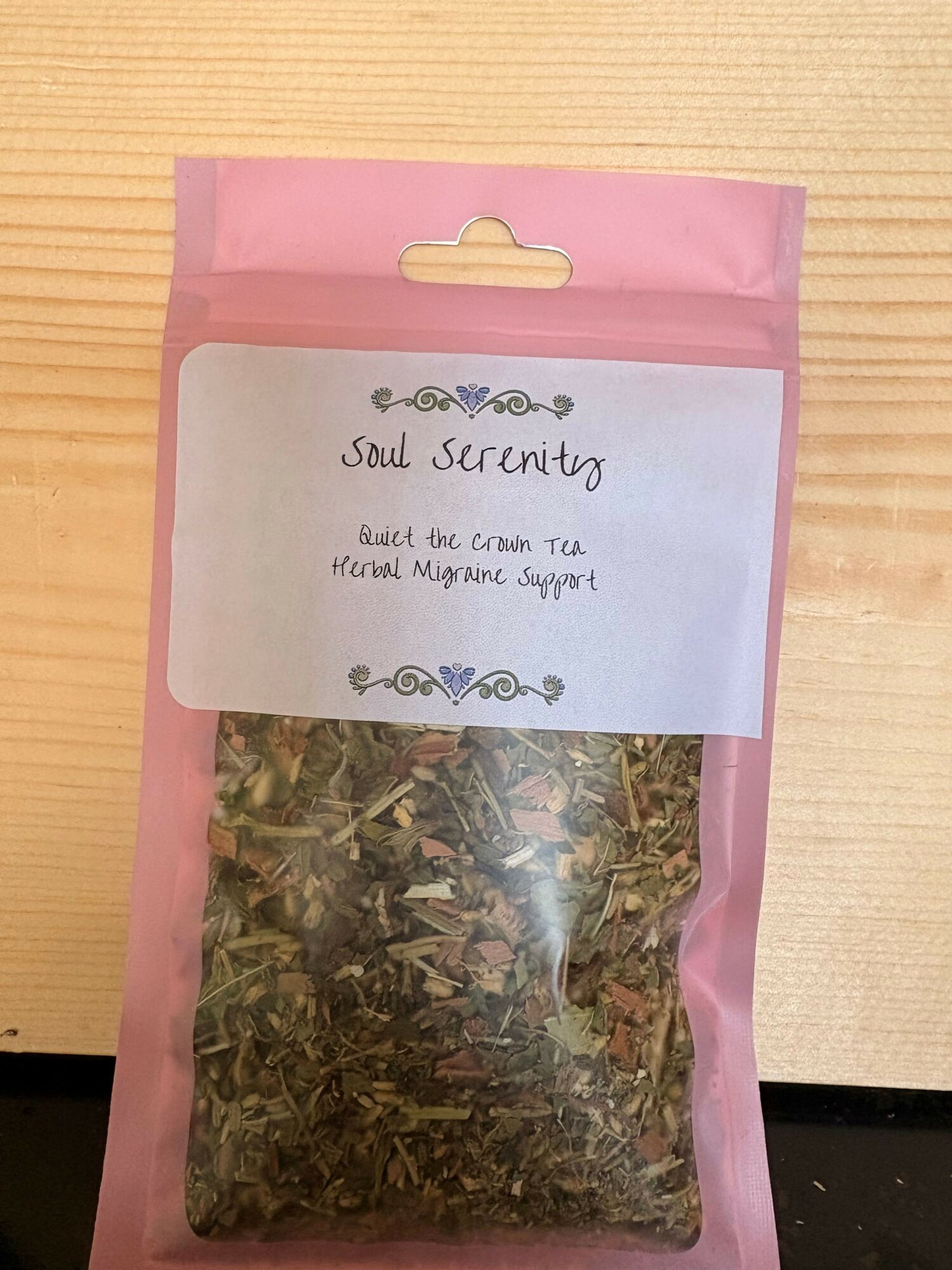 Pink package with a white label containing handwritten text and a transparent window showing dried herbs inside.