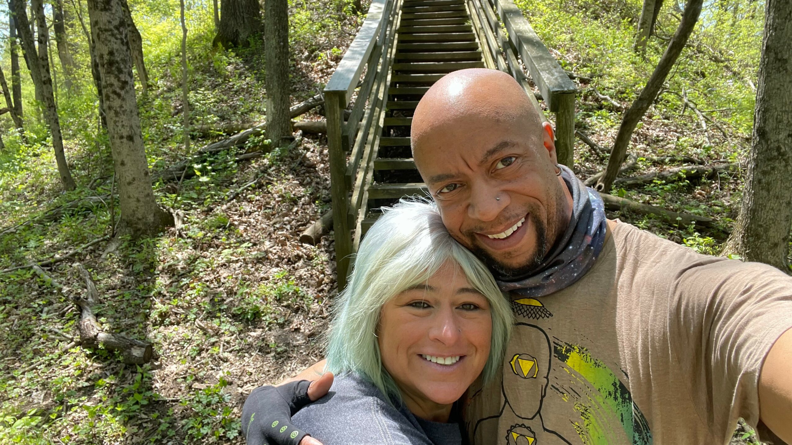 Two people smiling and taking a selfie on a wooden bridge in a forest.