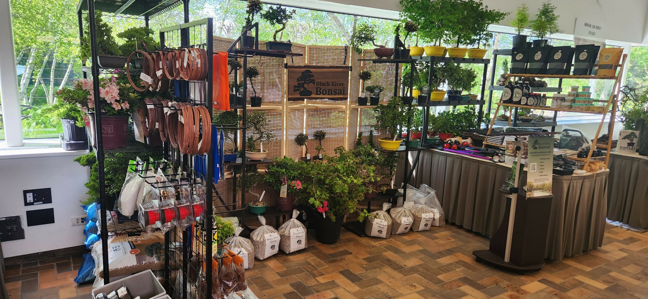 Indoor display of plants, pots, and gardening supplies on shelves and tables, with large windows and natural light.