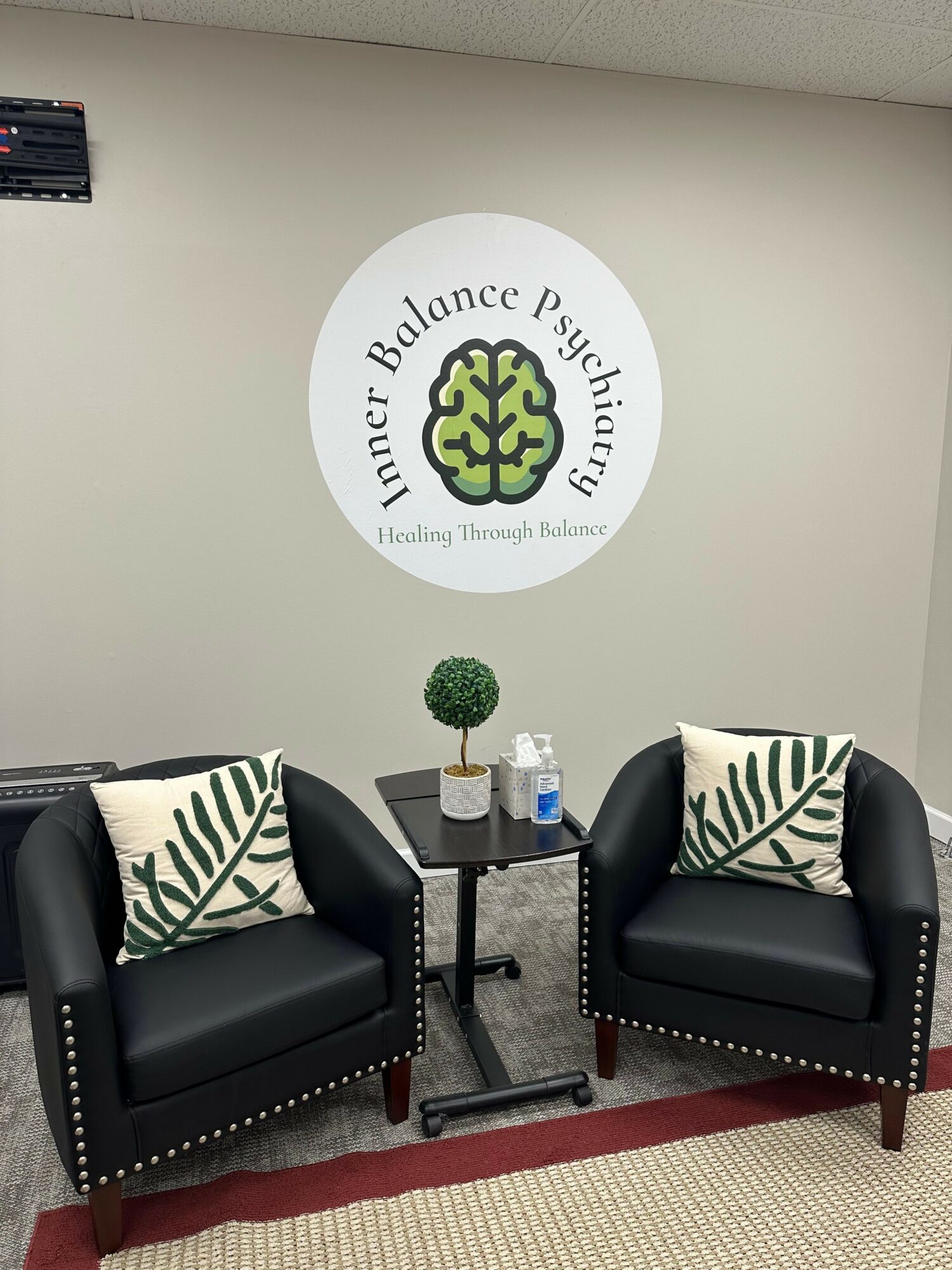Two black armchairs with patterned pillows face each other in a waiting area, with a small table and logo on wall.