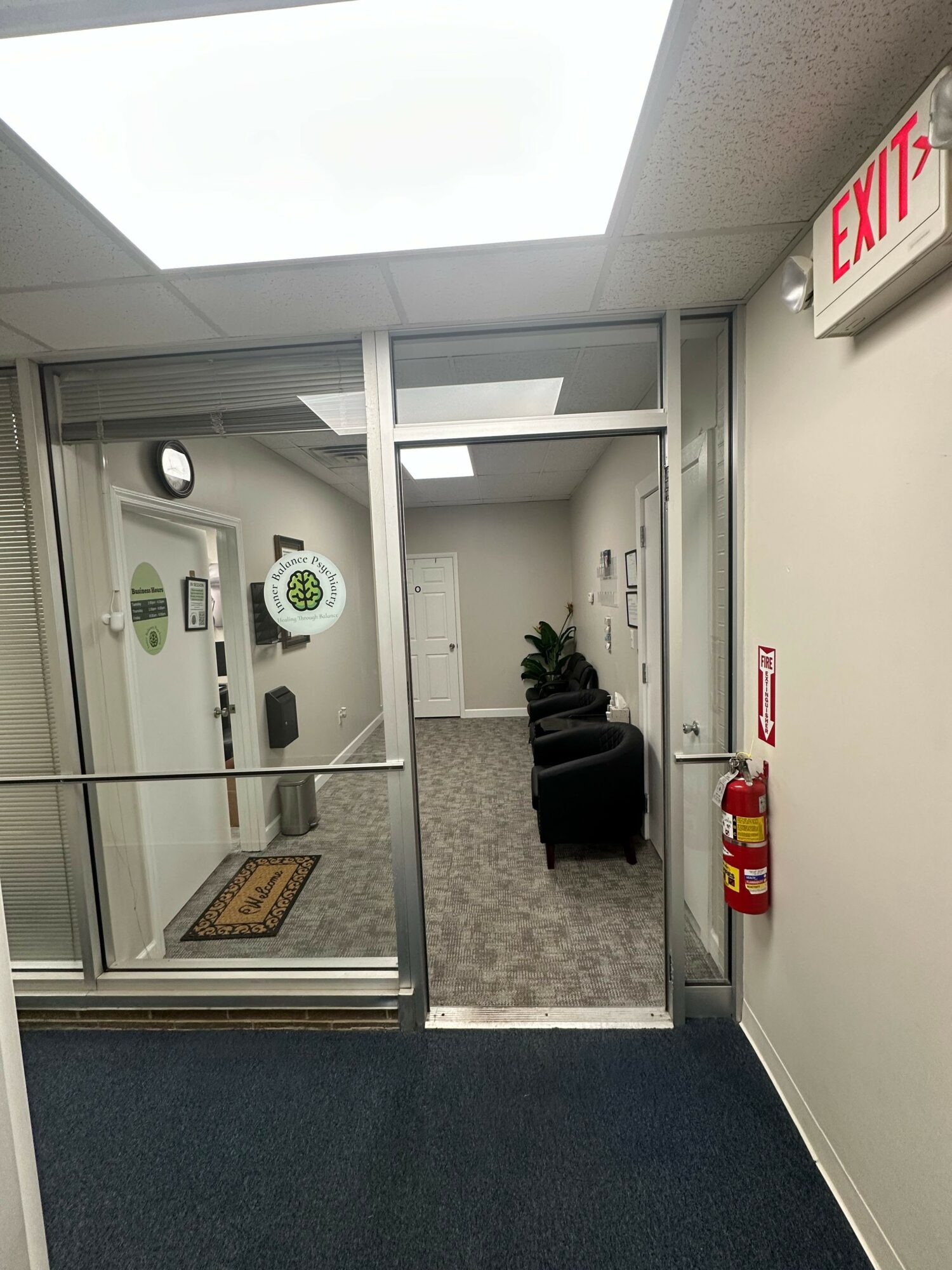 View through glass door into a waiting area with chairs, plants, and doorways, with an exit sign and fire extinguisher nearby.