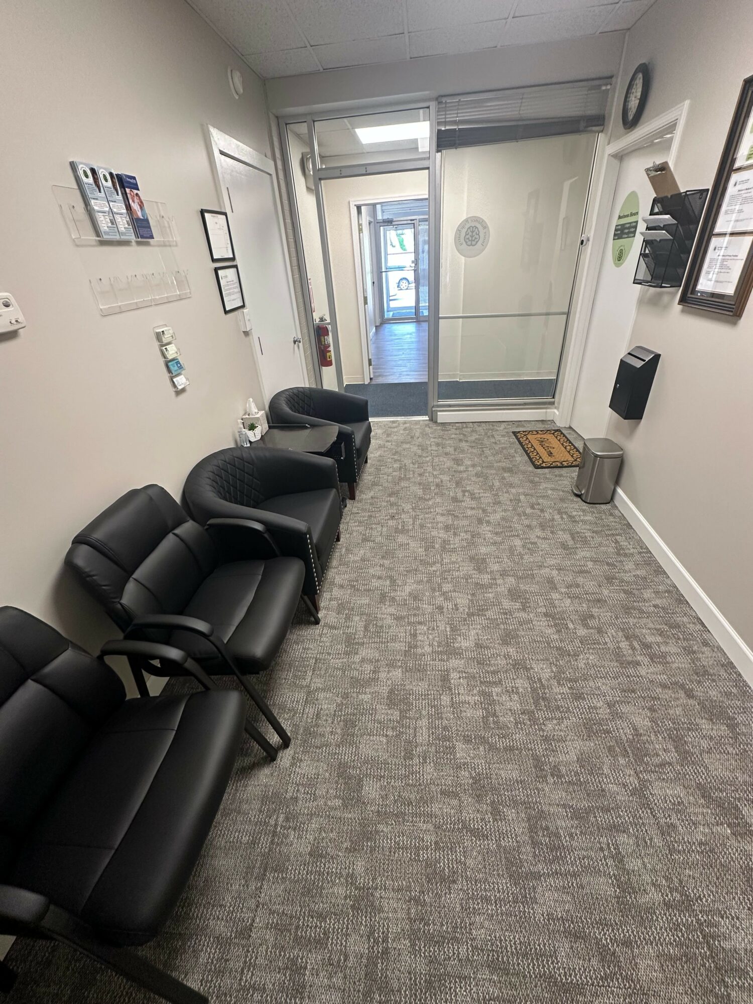 Waiting area with three black chairs, framed pictures on wall, glass door at end, carpeted floor, and a trash bin.