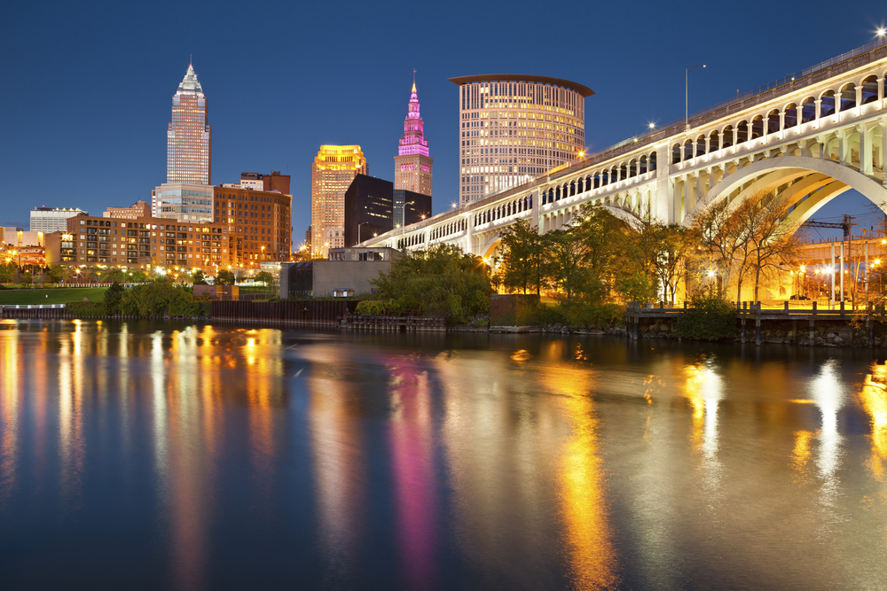City skyline at night with illuminated buildings, bridge, and reflections on water.