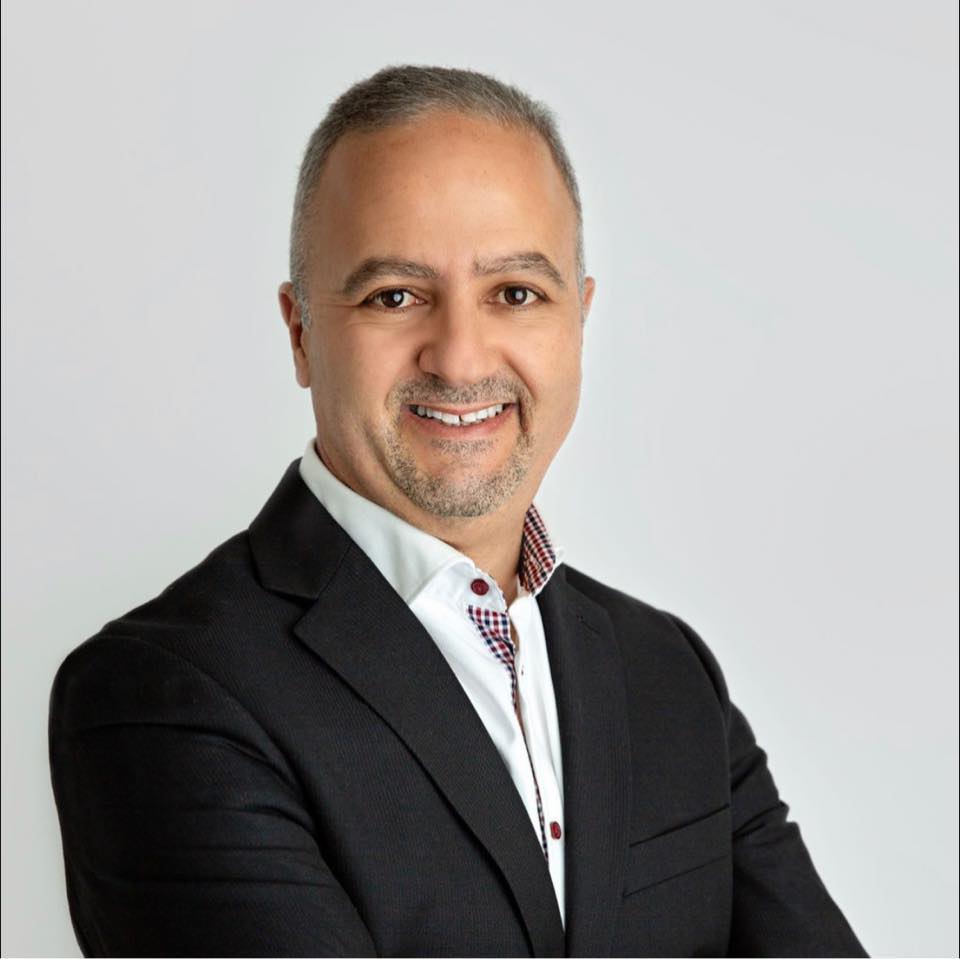 Man in a black blazer and white shirt smiling with arms crossed against a plain background.