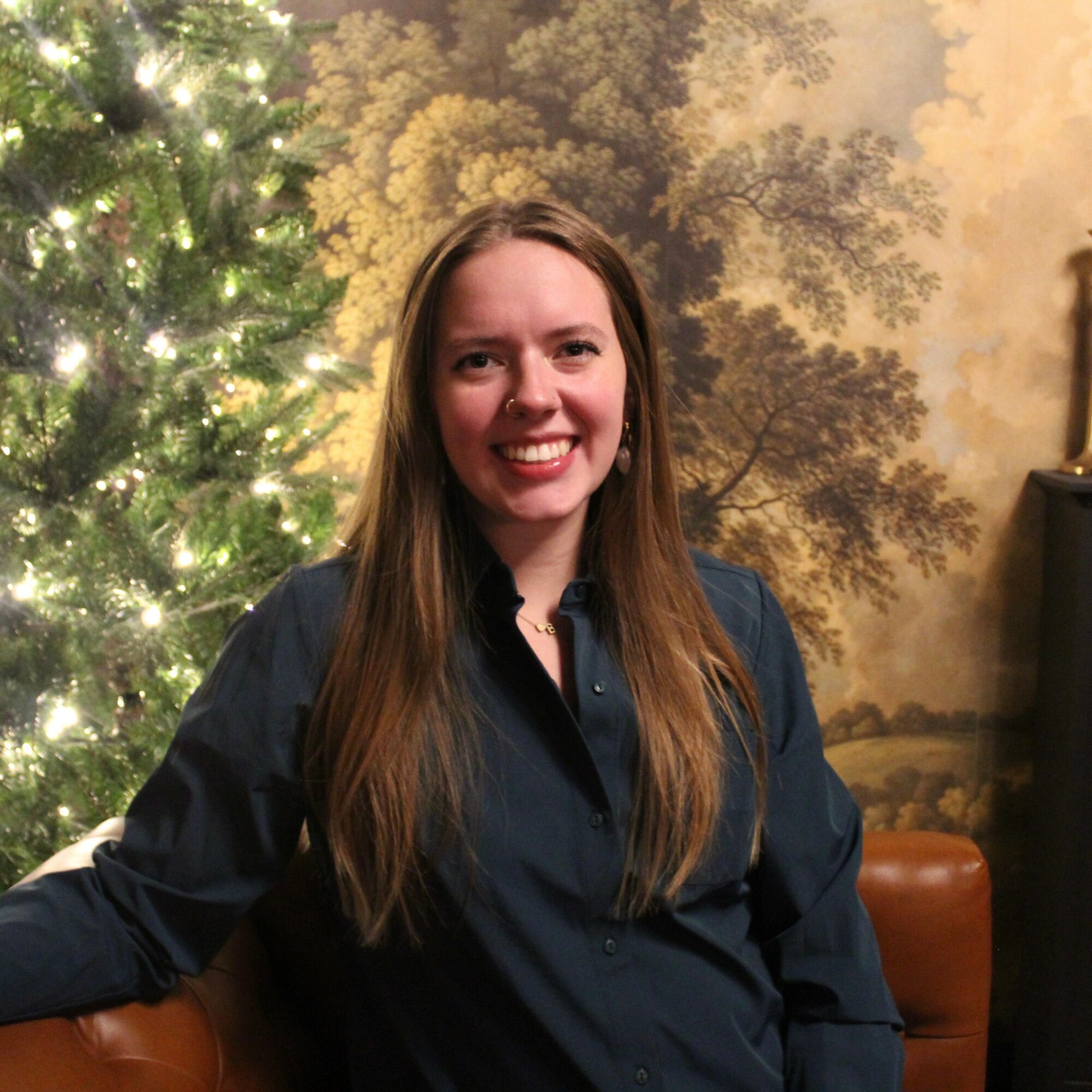 Young woman with long hair smiling, wearing a dark shirt, standing indoors near a decorated Christmas tree.