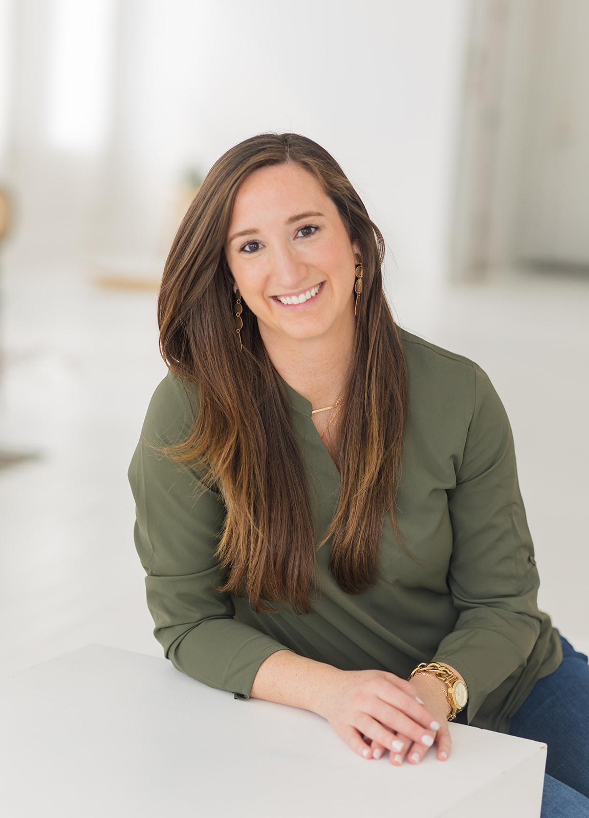 Young woman with long brown hair smiling, wearing a green top, sitting at a white surface in a bright room.