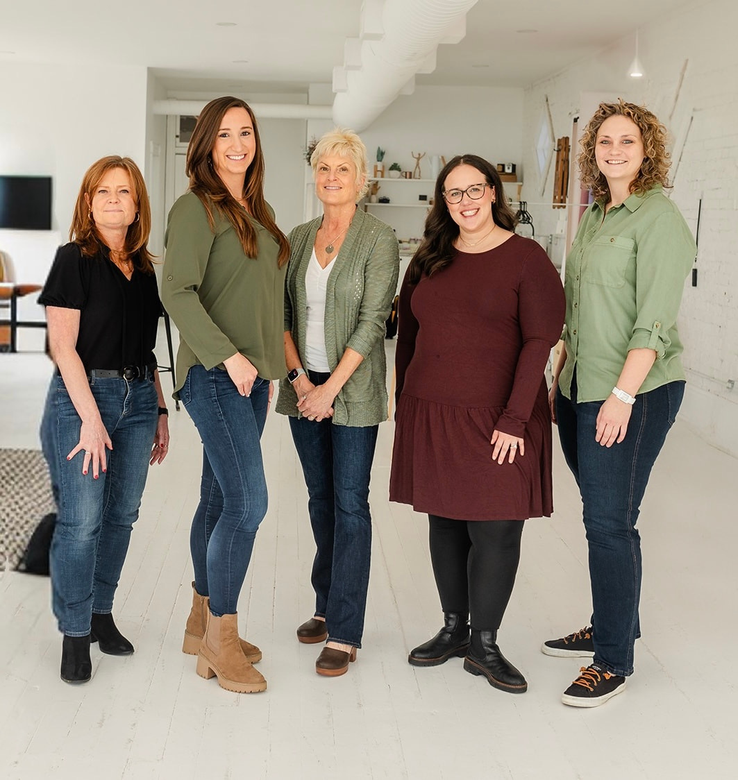 Five women standing together indoors, smiling, in casual clothing, with a white background and modern interior.