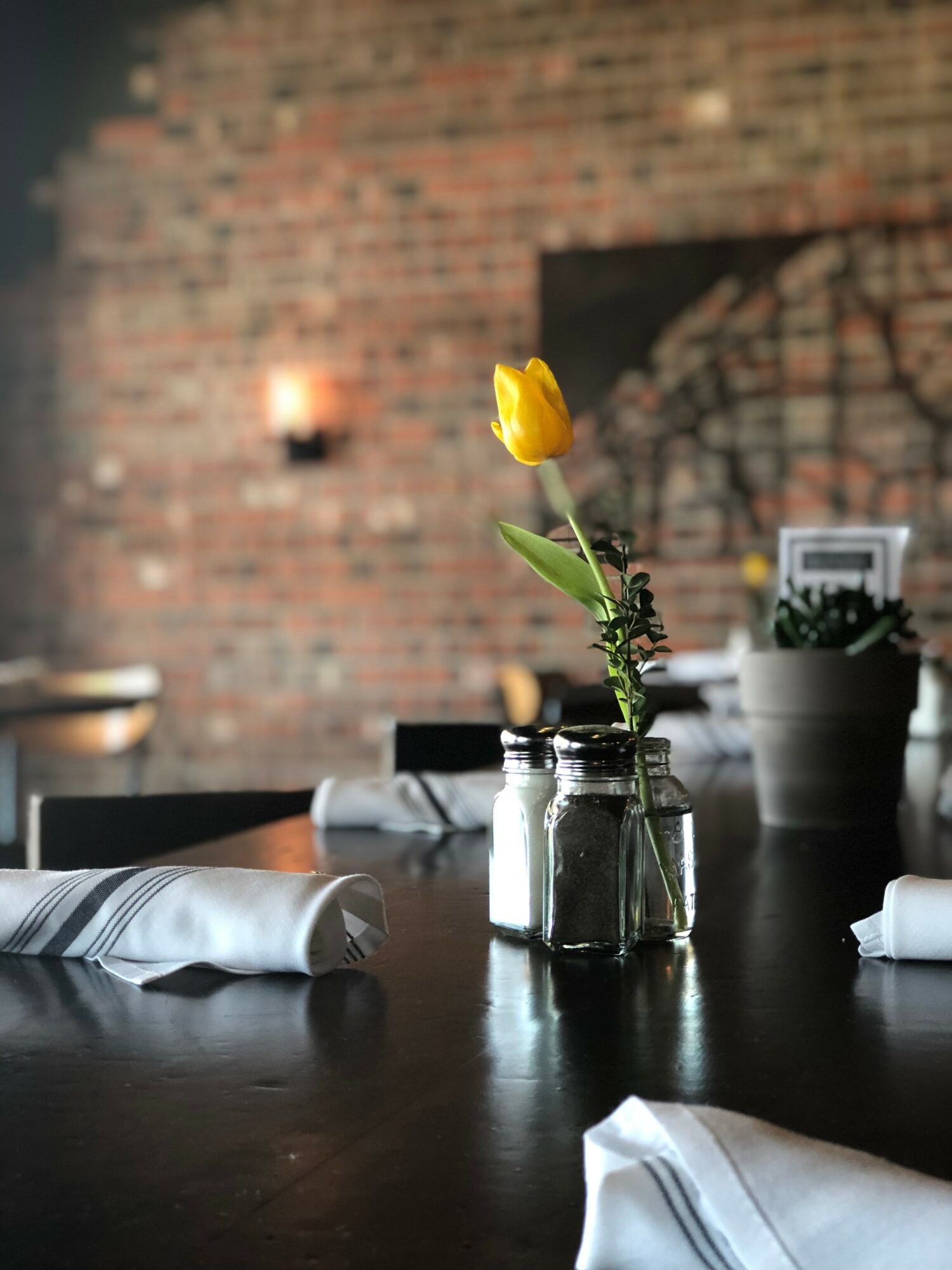 Yellow tulip in a glass jar on a dark table with a brick wall background.