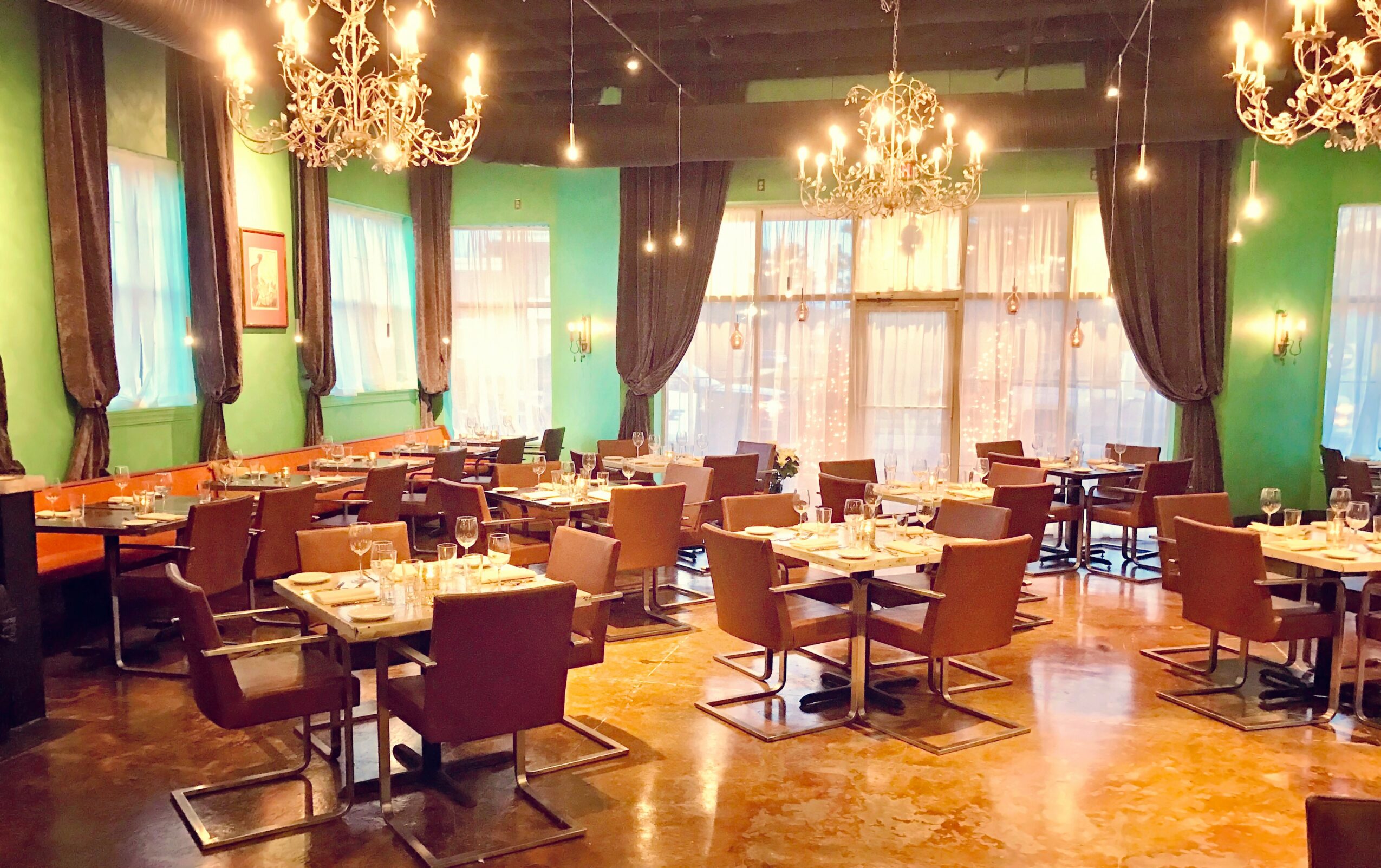 Elegant restaurant interior with green walls, chandeliers, large windows, and neatly arranged tables and chairs.