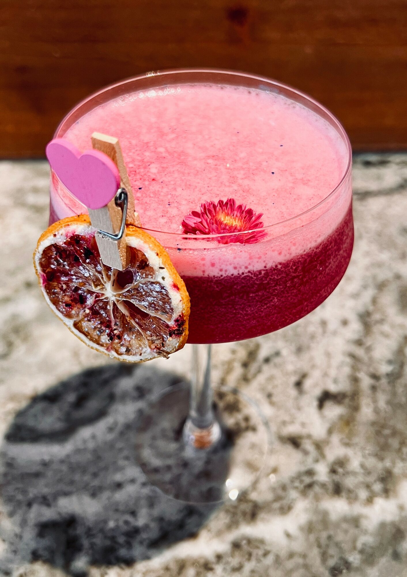 Pink cocktail in a glass with a dried citrus slice and a pink heart-shaped decoration, on a textured surface.