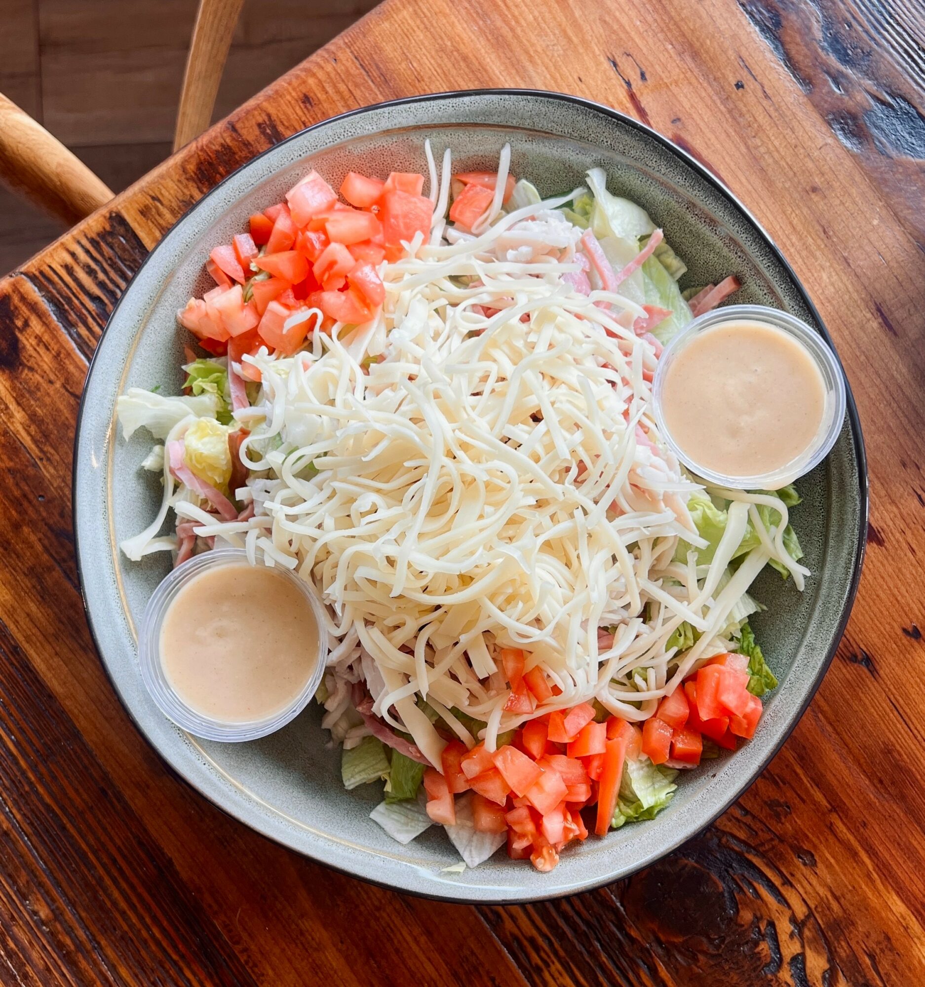 Bowl of salad with shredded cheese, chopped tomatoes, lettuce, and two small containers of dressing on a wooden table.