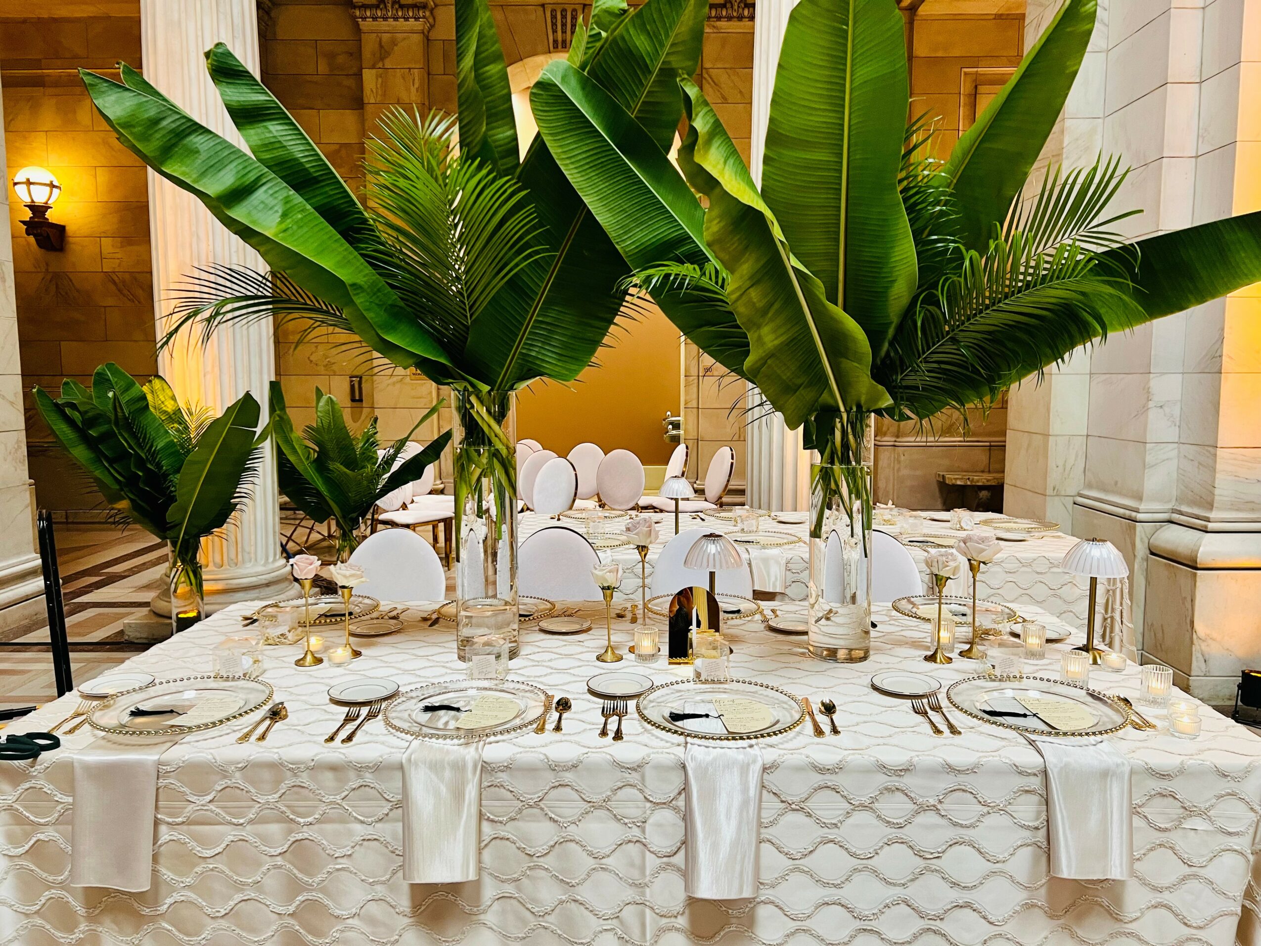 Elegant table setting with white tablecloth, plates, glasses, and cutlery, decorated with large green tropical plants in the background.