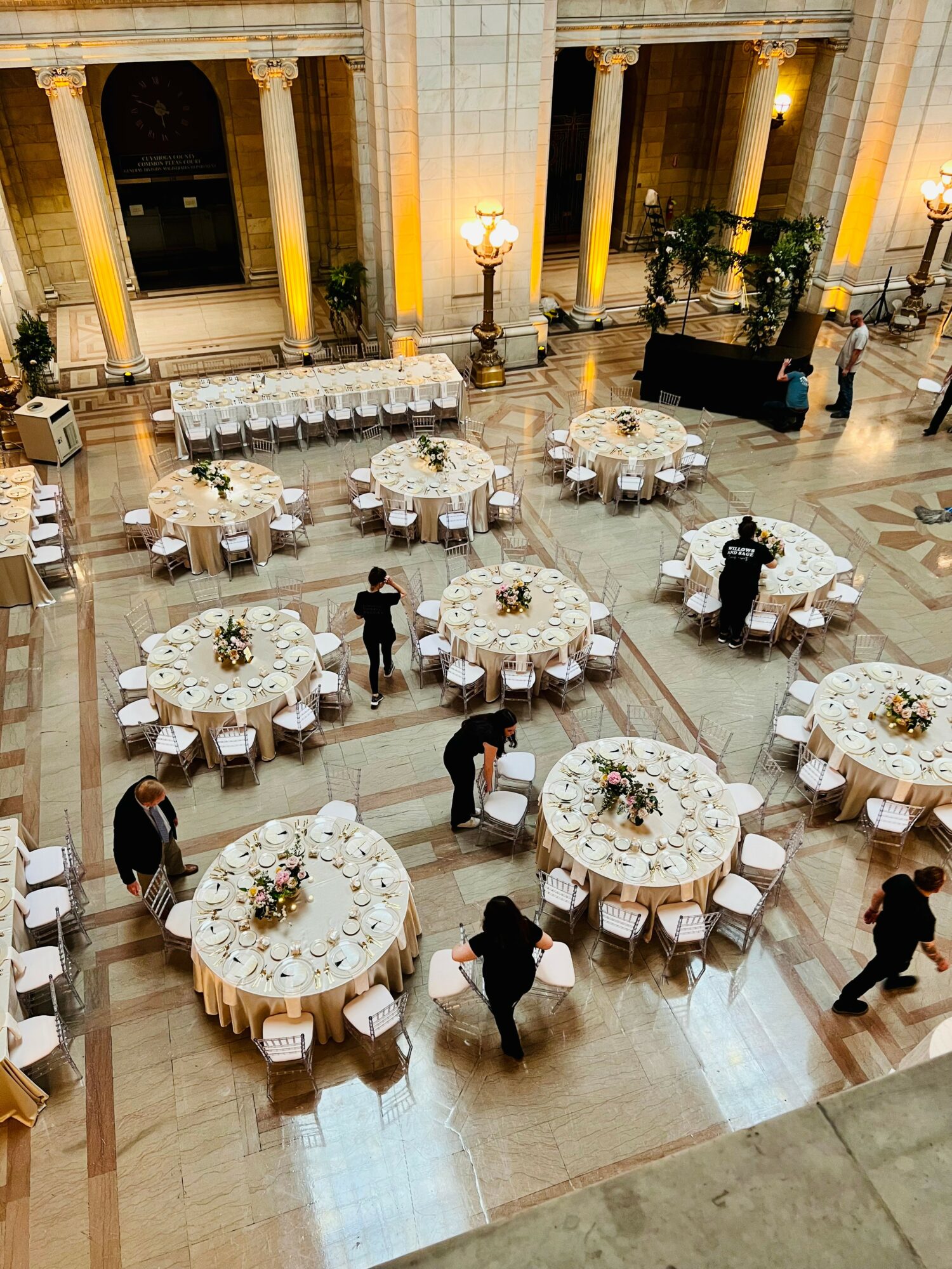 Elegant event with round tables, floral centerpieces, and people walking in a grand hall with tall columns and warm lighting.