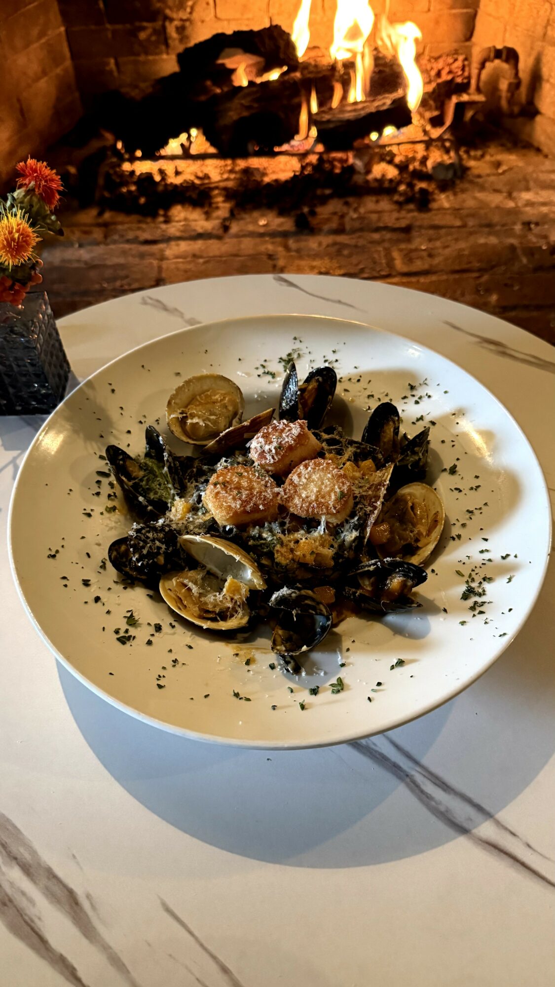 Plate of cooked mussels with herbs and seasoning on a white table, fireplace in background.