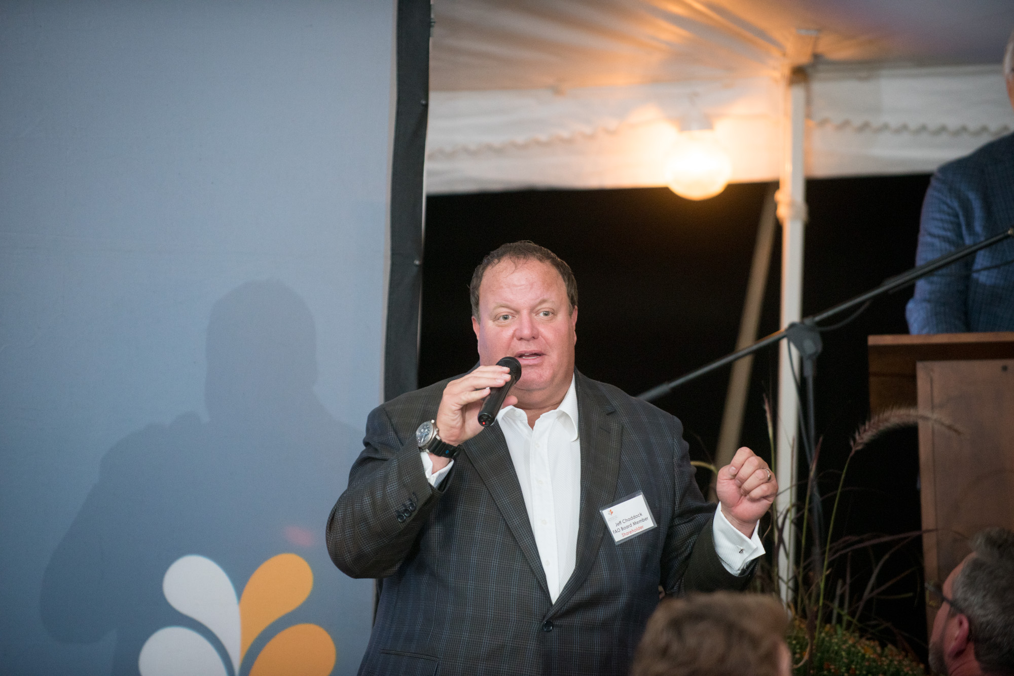 Man speaking into microphone at event, wearing suit and white shirt, with audience in foreground.