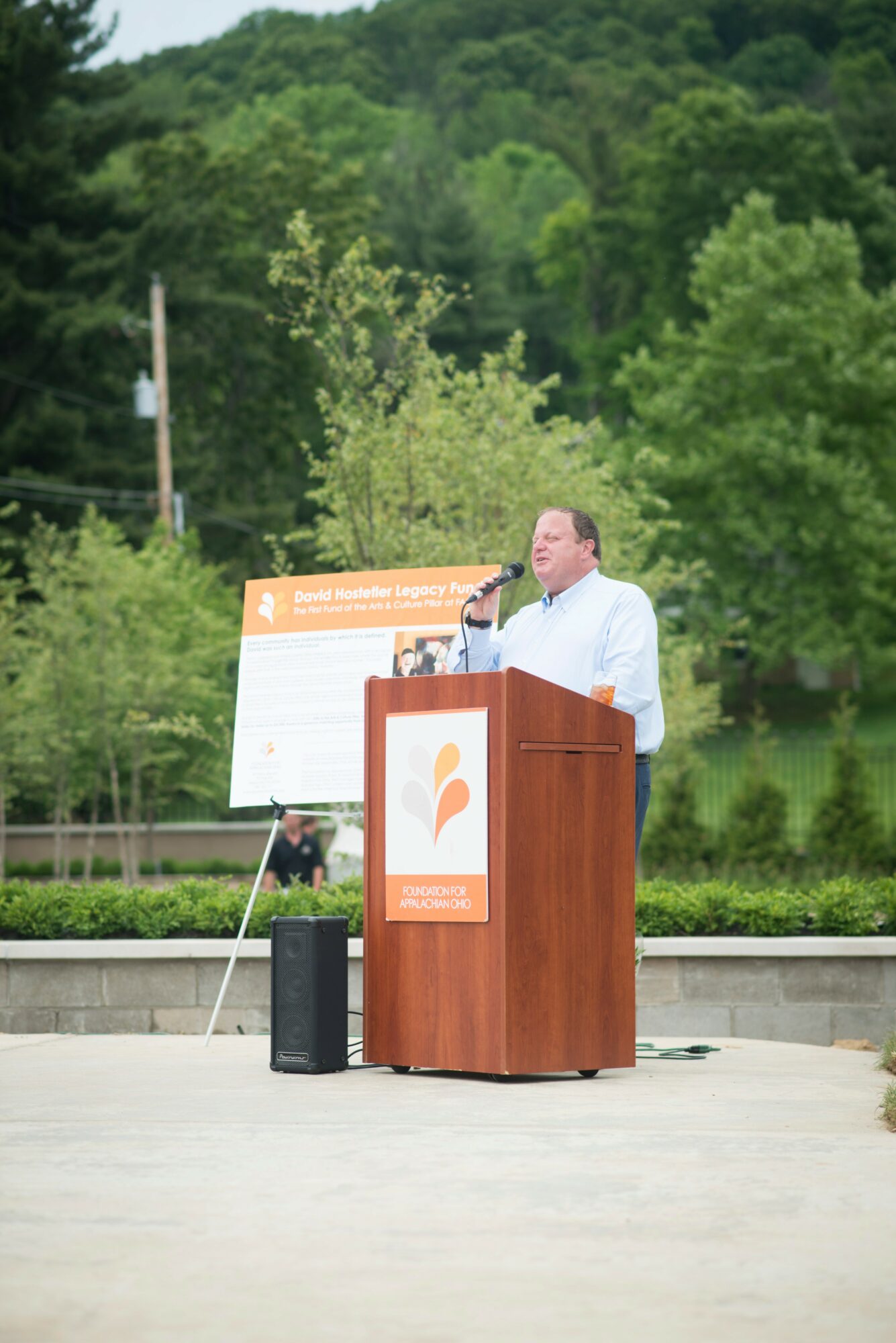 Man speaking at podium outdoors with trees and greenery in background.