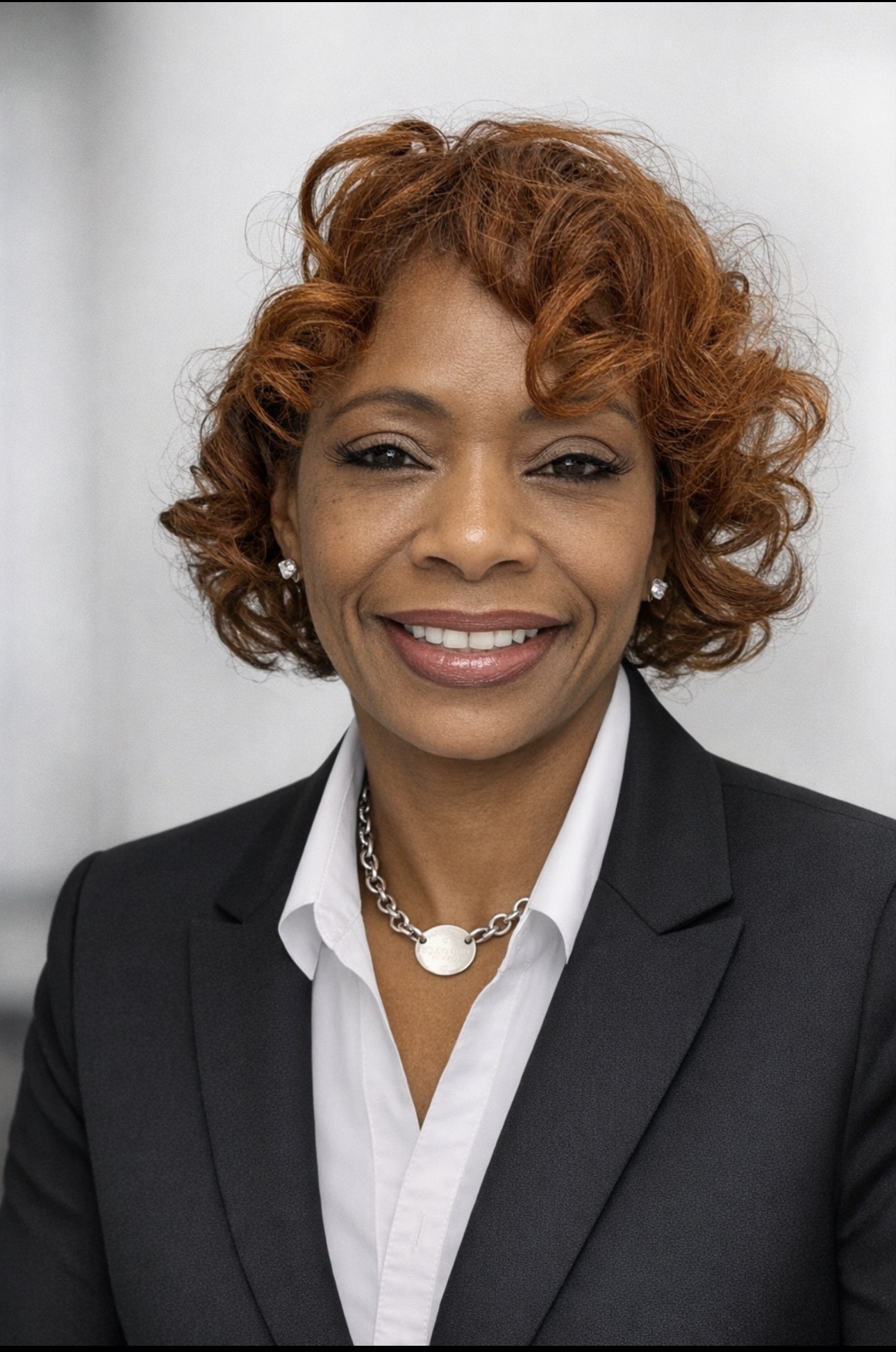 Woman with curly red hair wearing a black blazer and white shirt, smiling, against a light background.