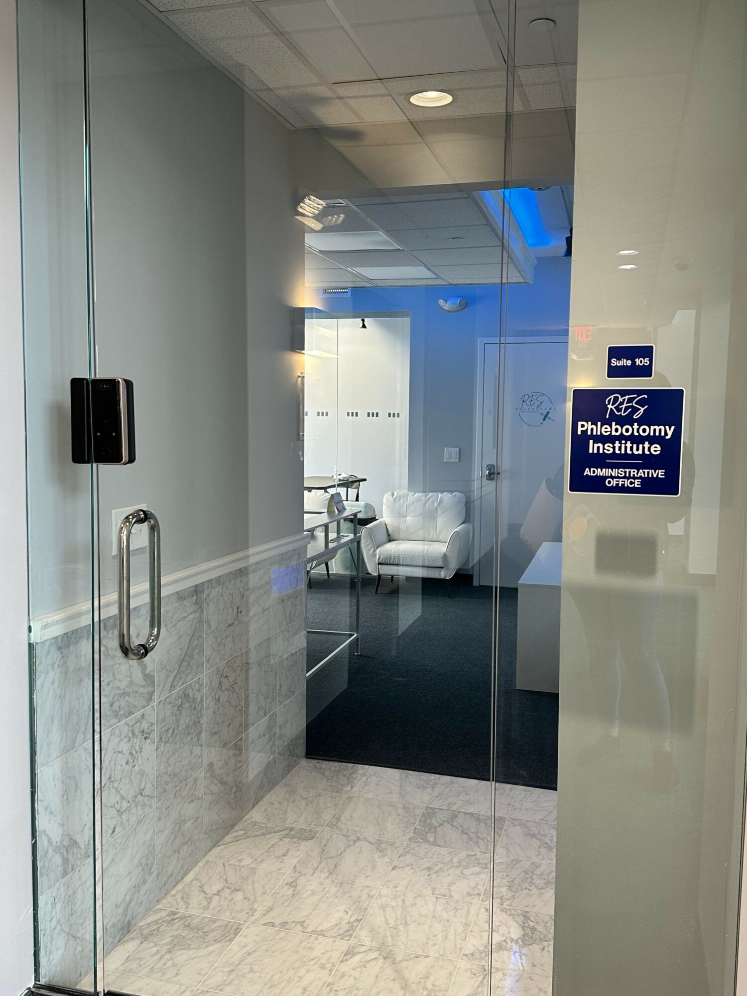 View through glass door into a room with white chairs and a table, blue ceiling light, and a blue sign on the wall.