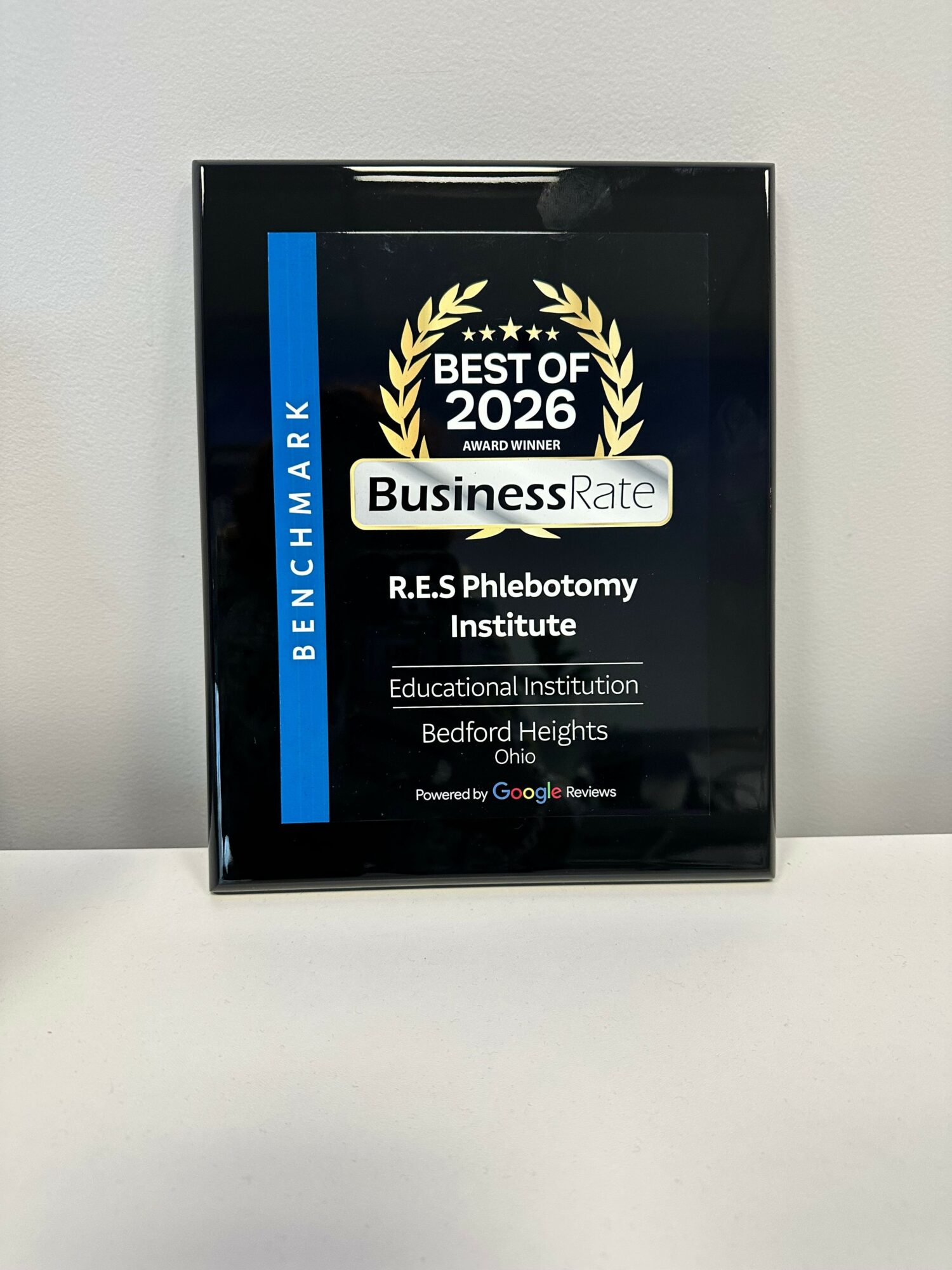 Framed award plaque with black background, gold laurel, and text, mounted on a white wall.