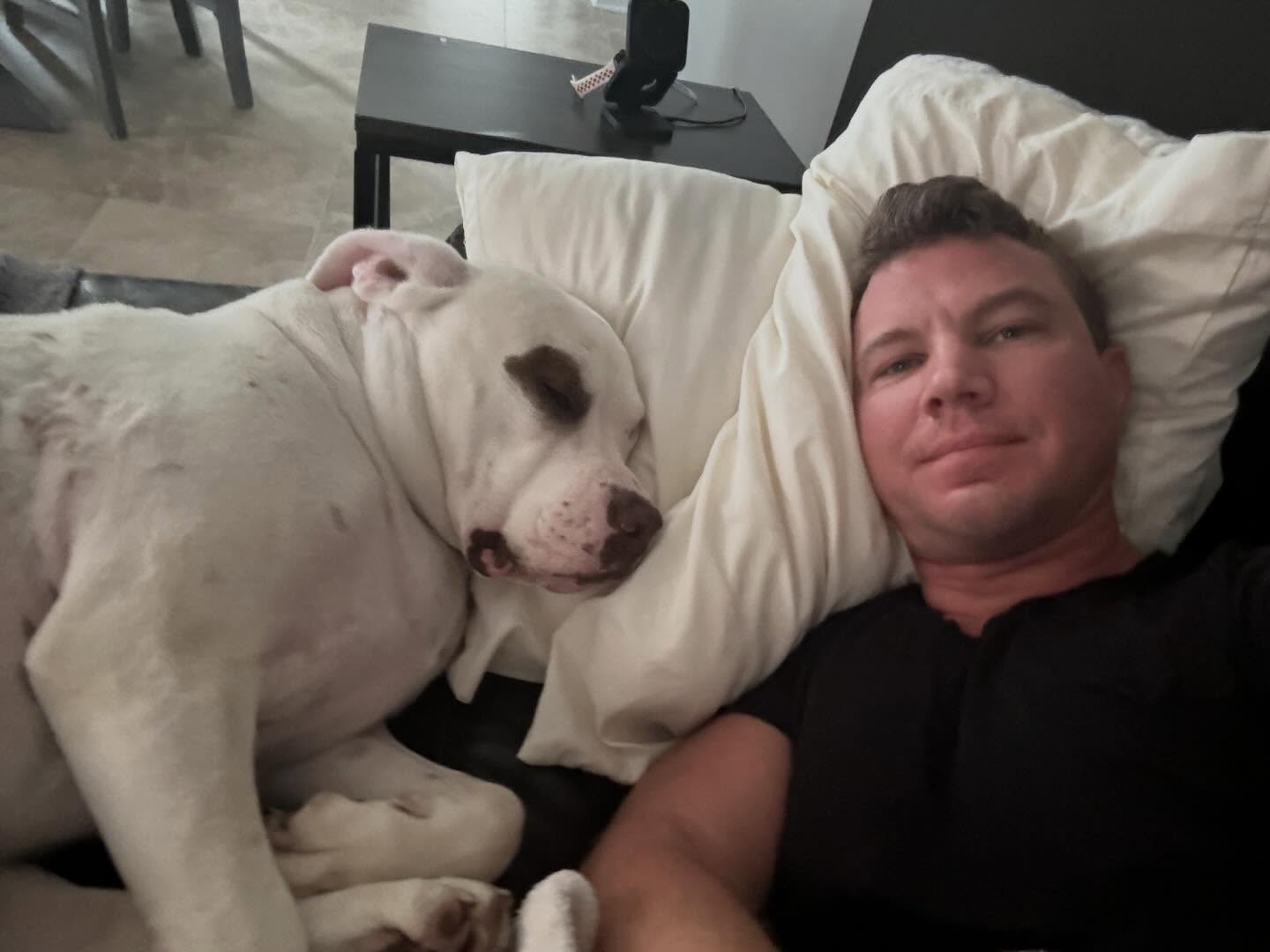 Person lying on a bed with a large white dog resting its head on a pillow beside them.
