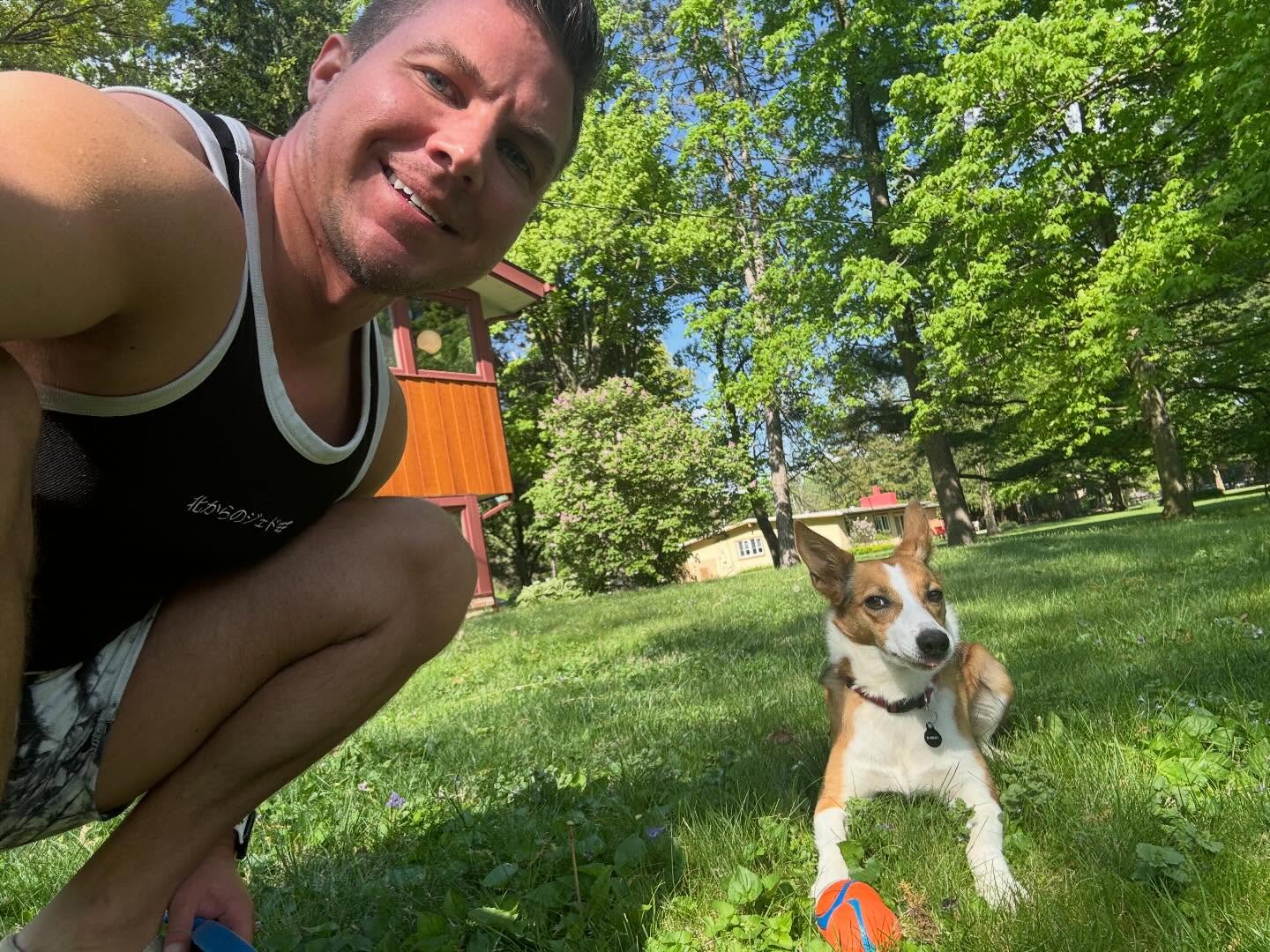 Man smiling outdoors with a dog on grass, trees and a building in background.