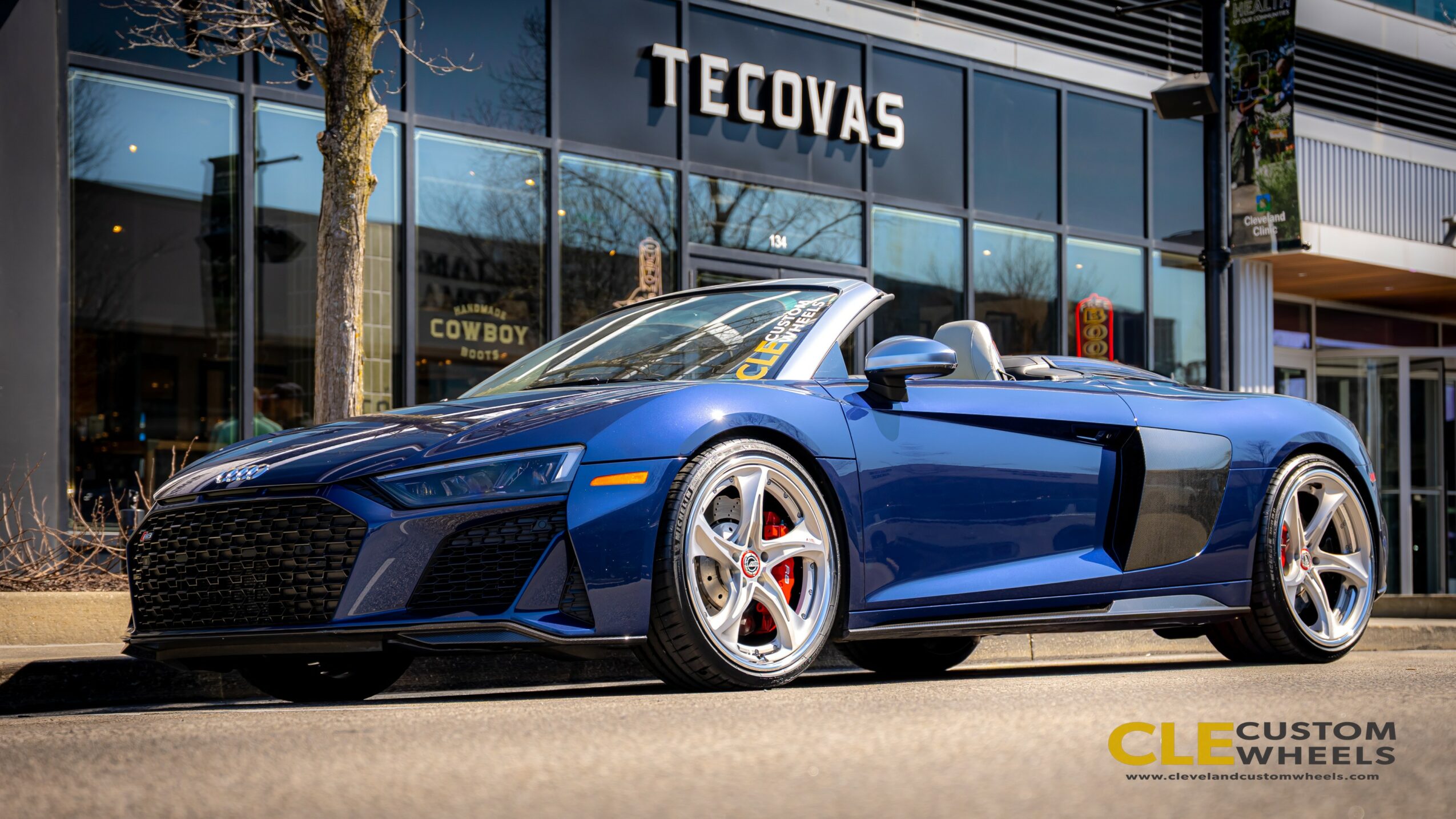 Blue convertible sports car parked in front of a Tecovas store with glass windows and signage.