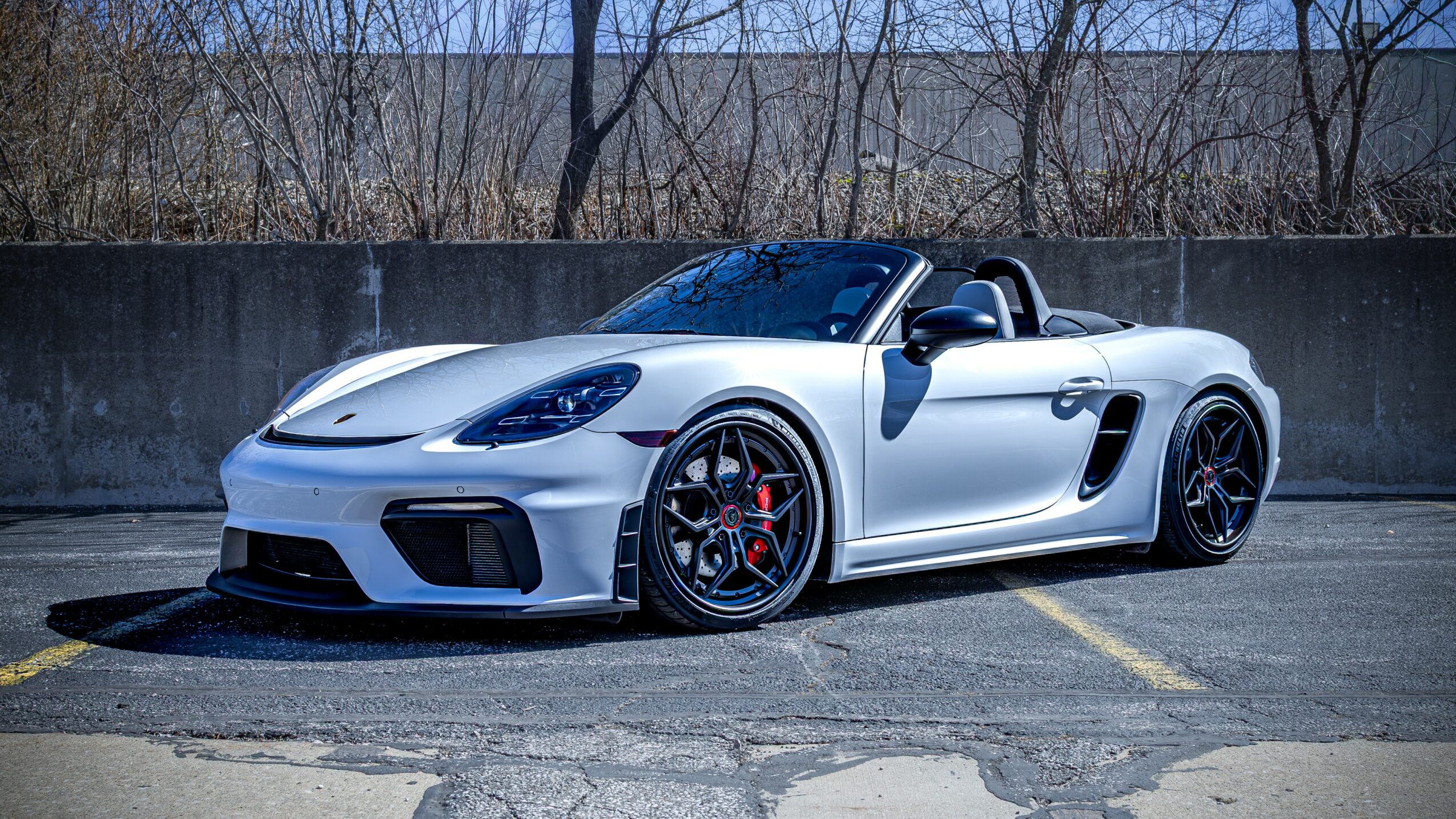 White convertible sports car parked on a paved lot with a concrete wall and trees in background.