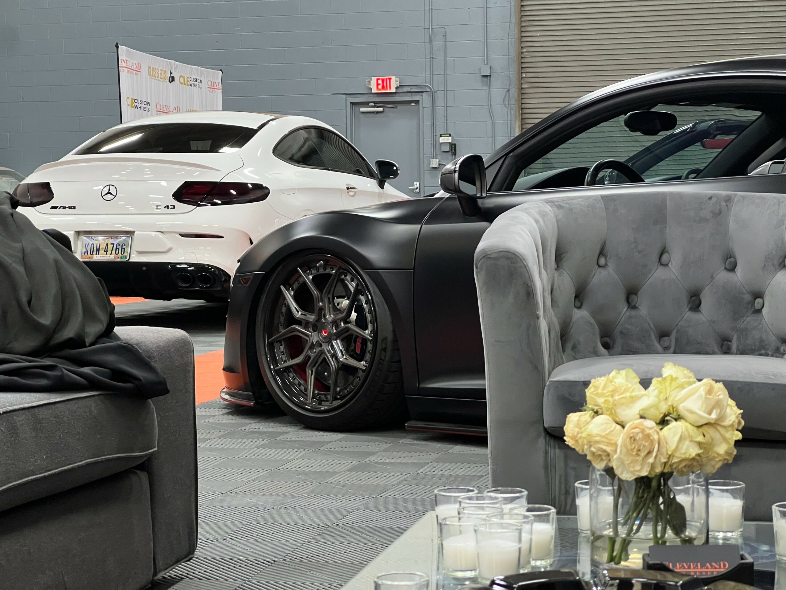 Two luxury cars parked indoors, one black and one white, with a gray sofa and a flower arrangement in the foreground.