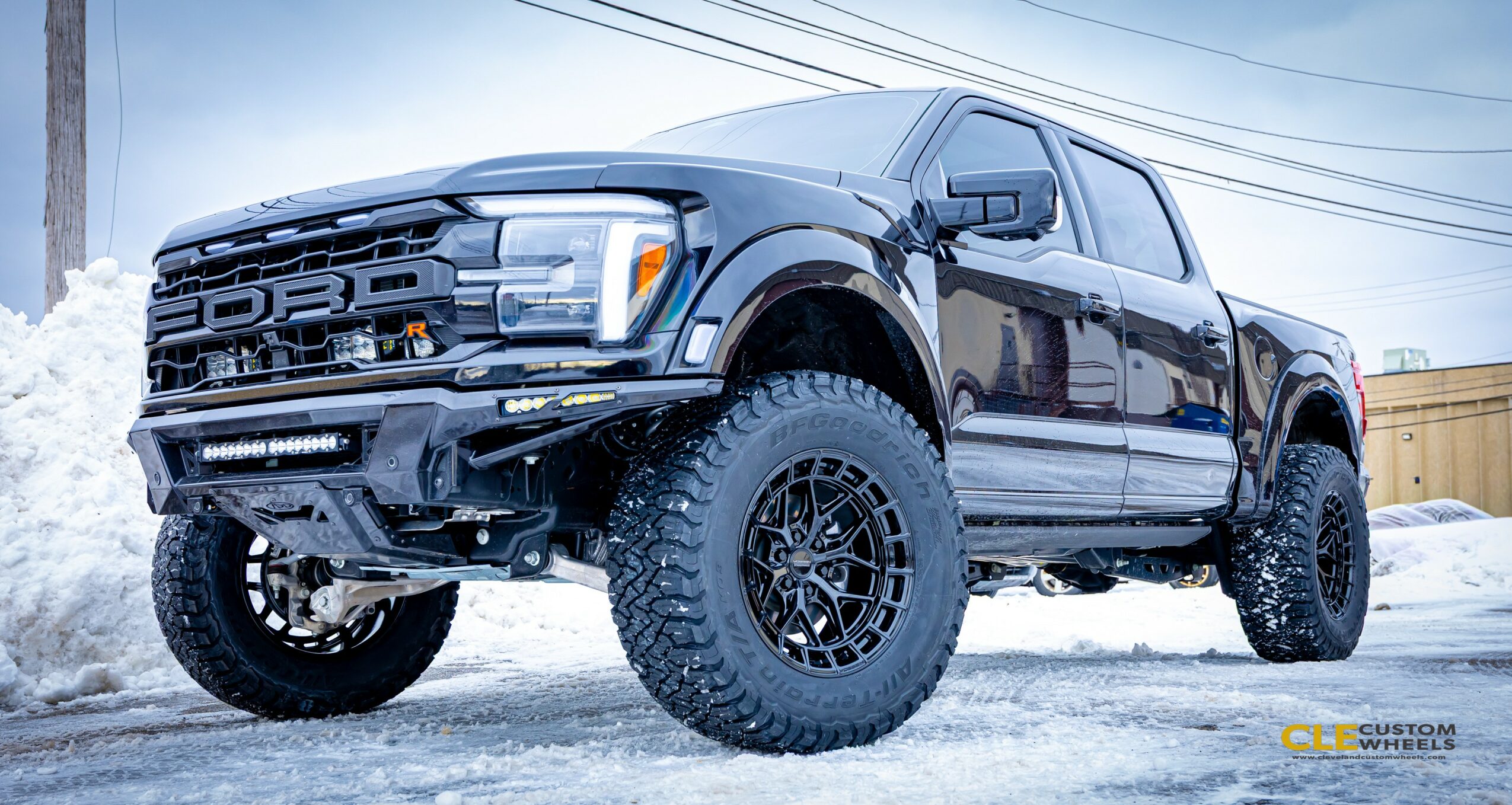 Black pickup truck with large tires parked on snowy ground, with snow and a building in background.
