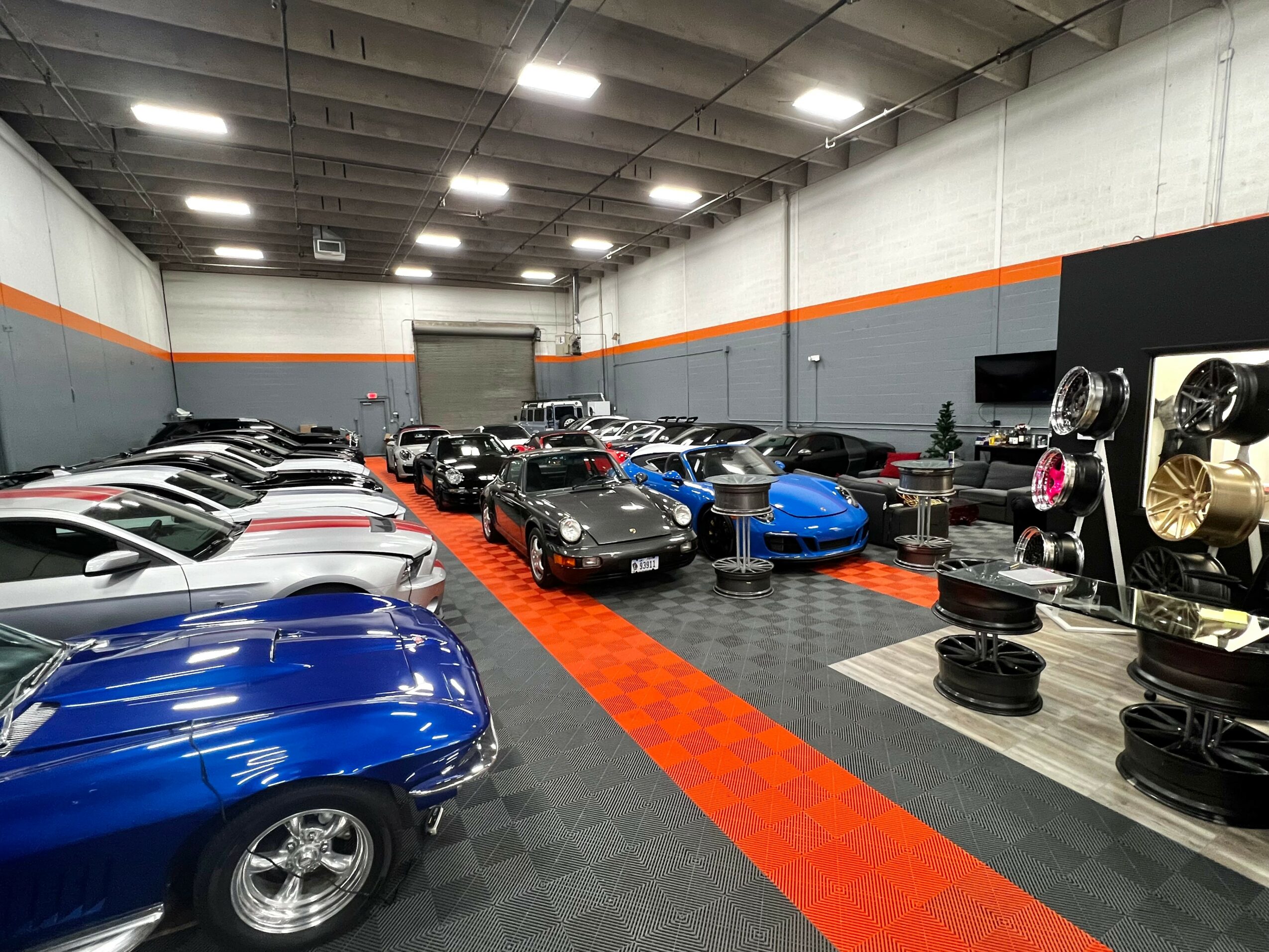Indoor car showroom with various classic and sports cars on display, black and gray flooring, and a black and orange wall.